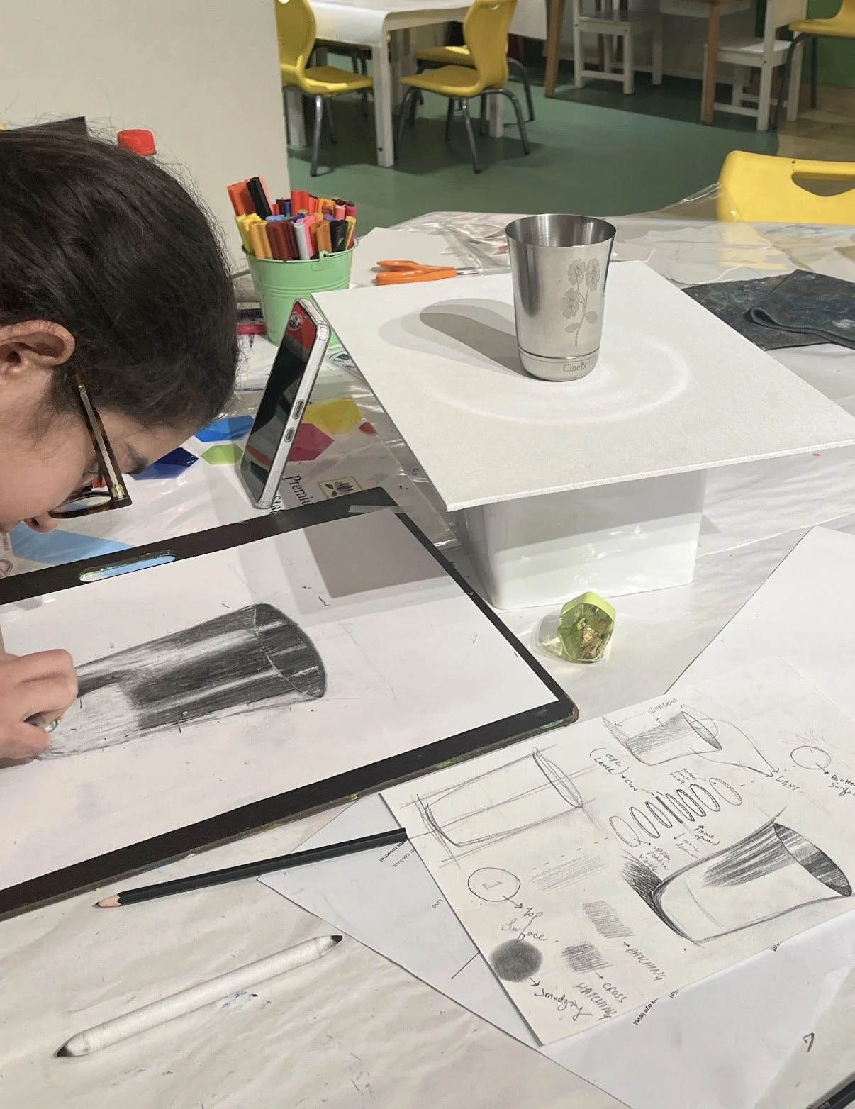 A person sketching a cylindrical object, possibly a glass, on paper with technical drawings and notes on the table. Various art supplies and a metal cup are nearby.
