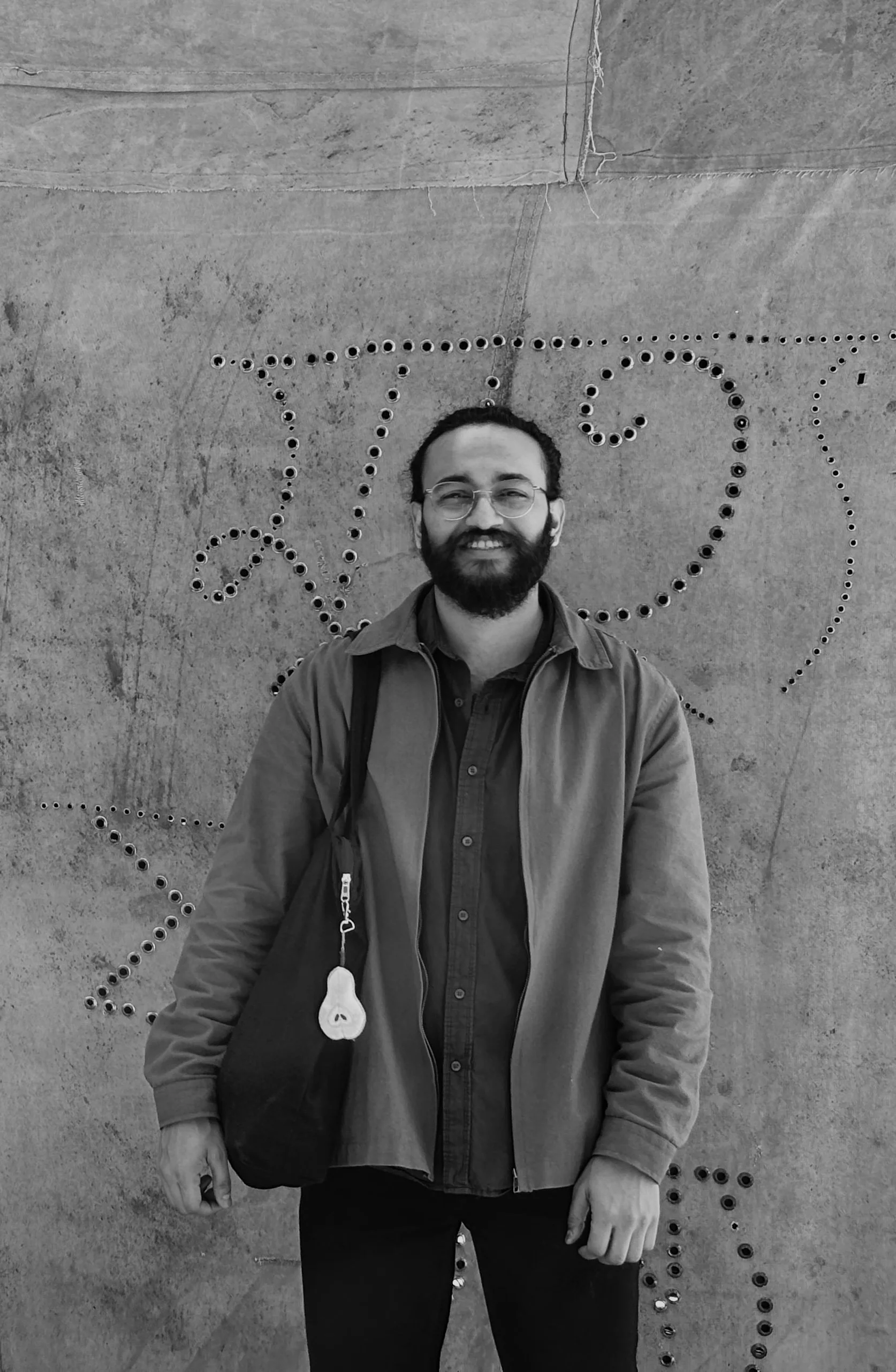 A smiling man with glasses and a beard standing in front of a concrete wall with decorative metallic dots arranged in a swirling pattern.