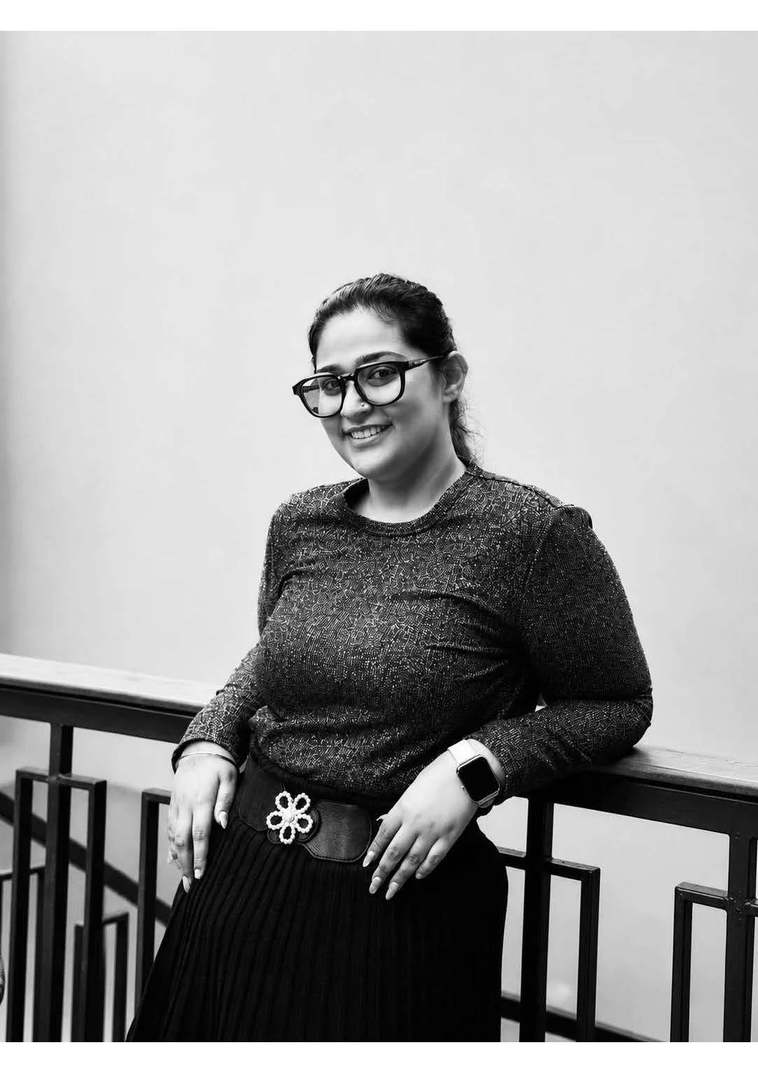 Black-and-white photo of a woman with glasses, wearing a glittery top, standing against a plain wall and leaning on a railing, smiling at the camera.