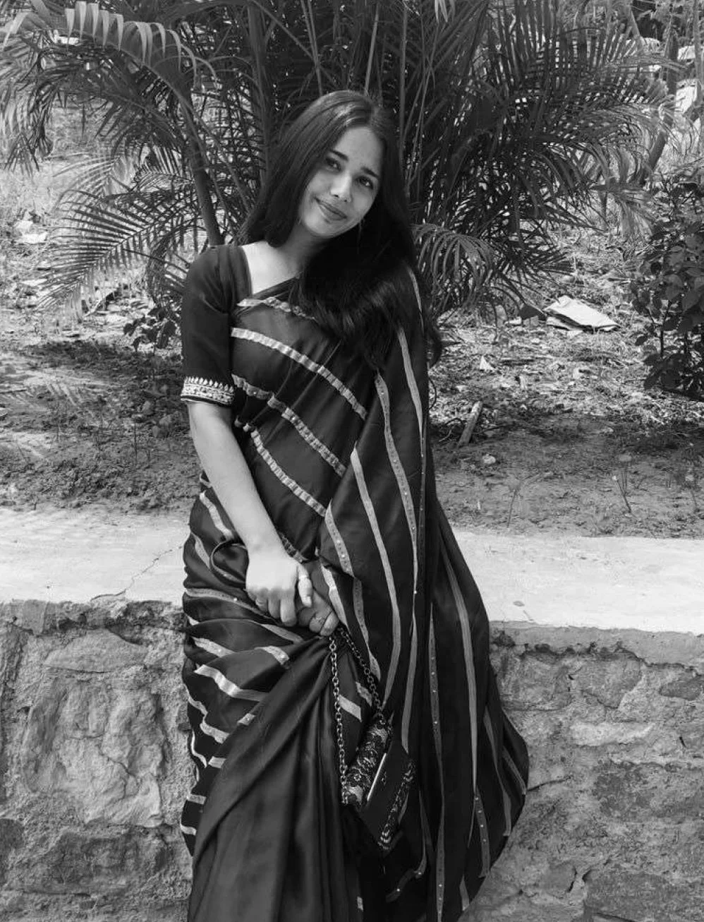 A young woman in a traditional sari, holding the edge of her sari with one hand and a small handbag with the other, standing outdoors in front of lush tropical plants.