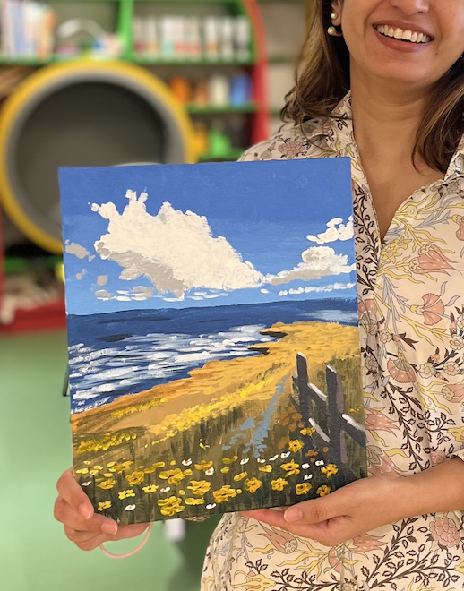 Woman holding a painting of a beach landscape with yellow flowers, a fence, ocean waves, and a partly cloudy sky.