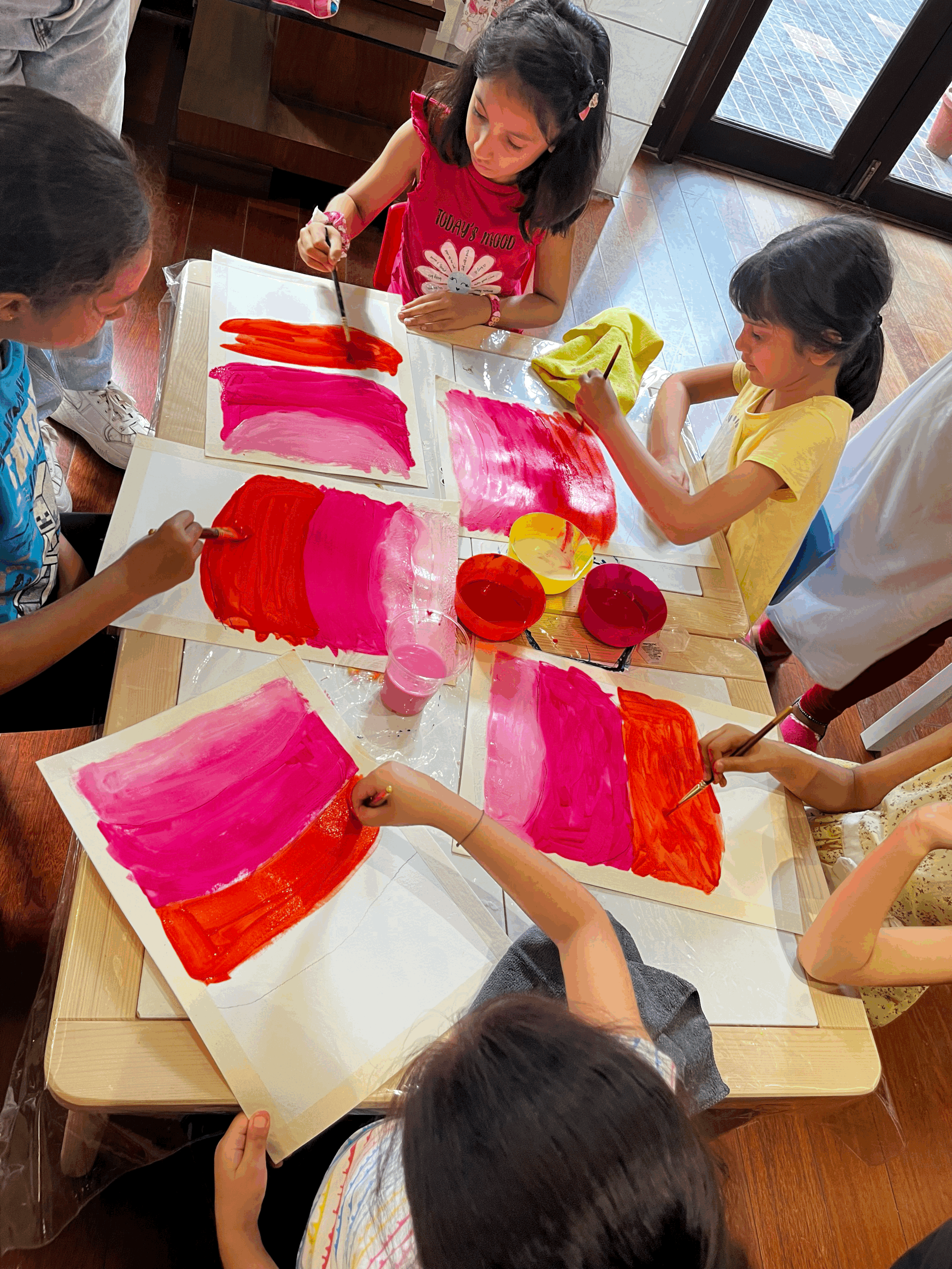 Children painting colorful abstract artwork with brushes on large sheets of paper, sitting around a wooden table indoors.