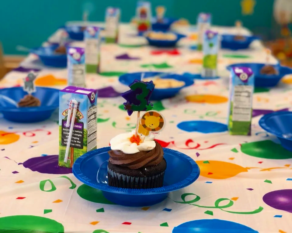 A table decorated with colorful balloons and streamers for a birthday party, featuring a cupcake with white and chocolate frosting, a lion topper, and a purple number two candle. Surrounding the cupcake are juice boxes and blue plates.