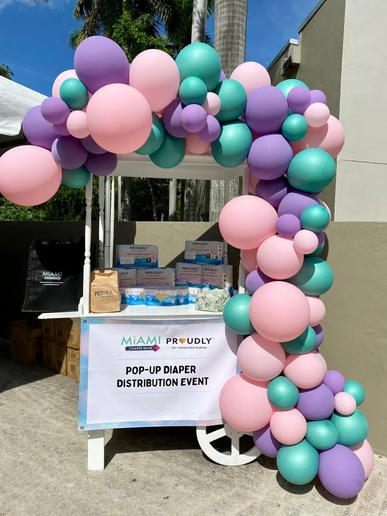 A mobile booth decorated with pastel pink, purple, and turquoise balloons, displaying a sign that says 'Miami Proudly Pop-Up Diaper Distribution Event'.