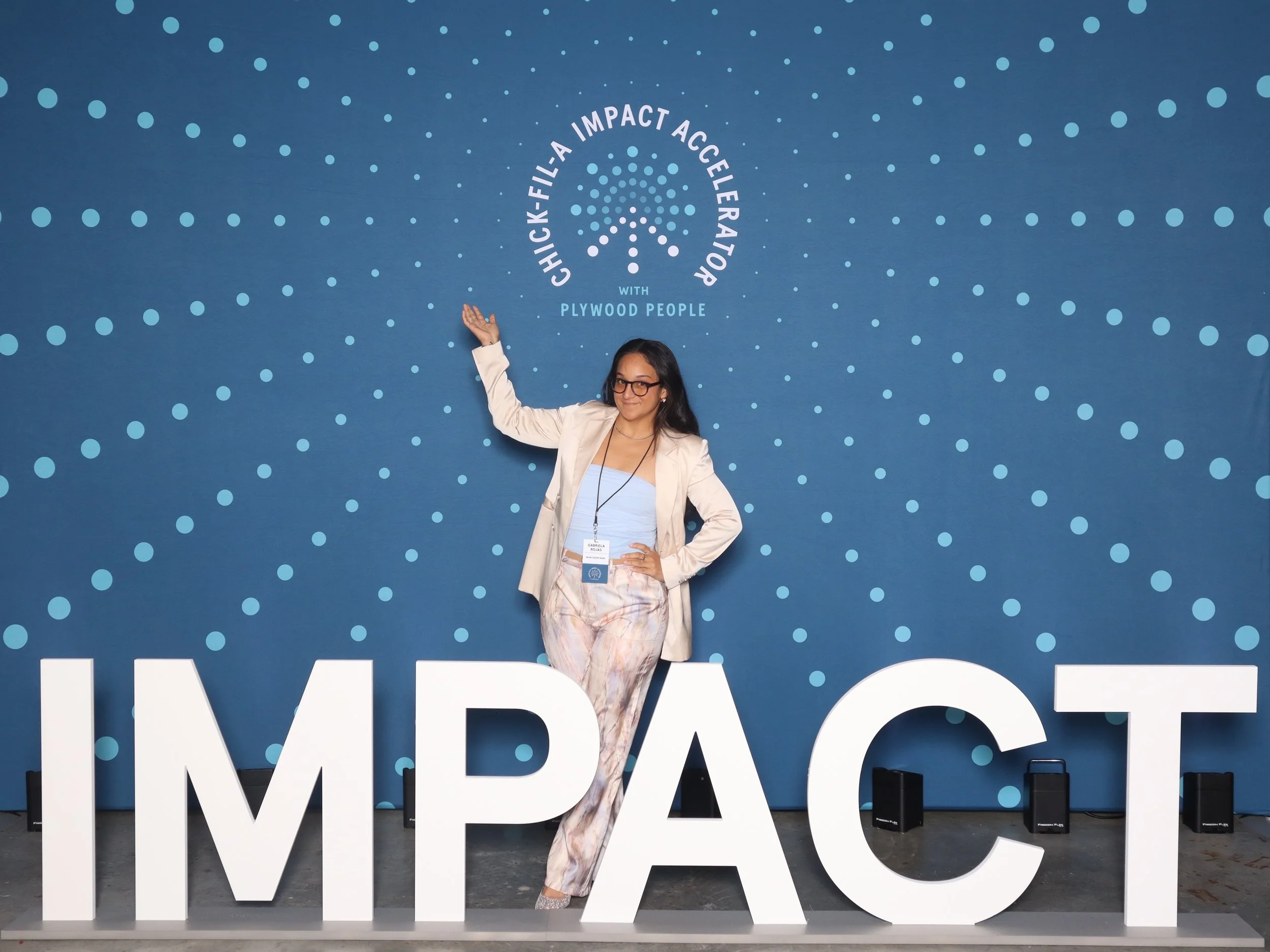 A woman standing in front of a blue backdrop with dotted patterns and text that says "Quick-Fil-A Impact Accelerator with Plywood People." She is dressed in a blazer and pastel-colored pants, and is posing with one hand on her hip and the other pointing upward.