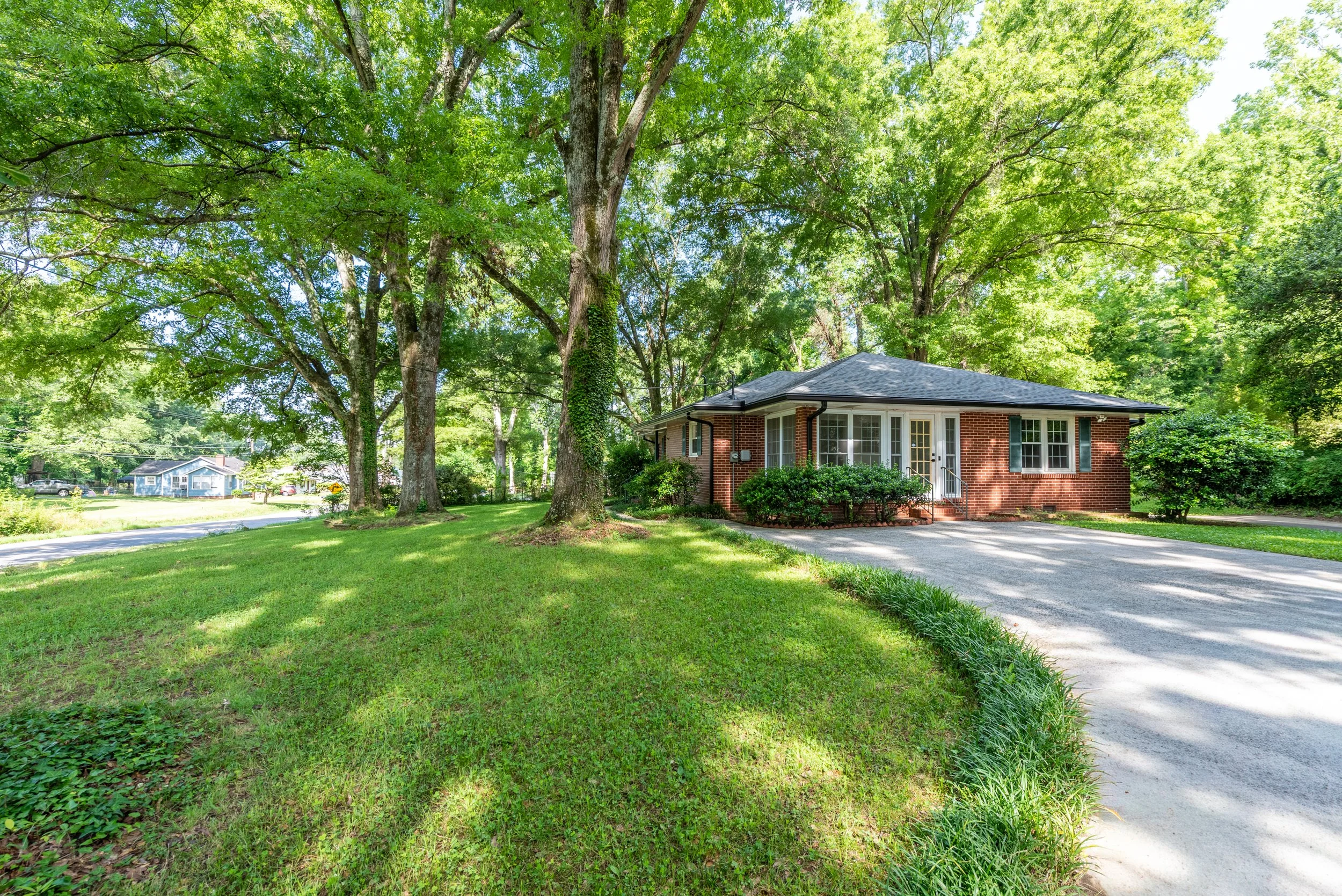 A single-story brick house with a gray roof, situated among tall green trees and a green lawn. There is a driveway in front of the house and smaller houses visible in the background.