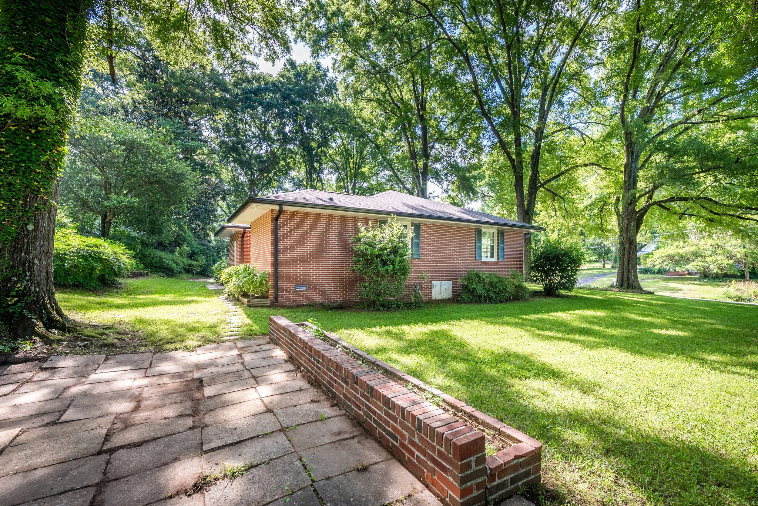 A brick house surrounded by tall trees and a well-maintained lawn, with a brick-lined sidewalk in the foreground.