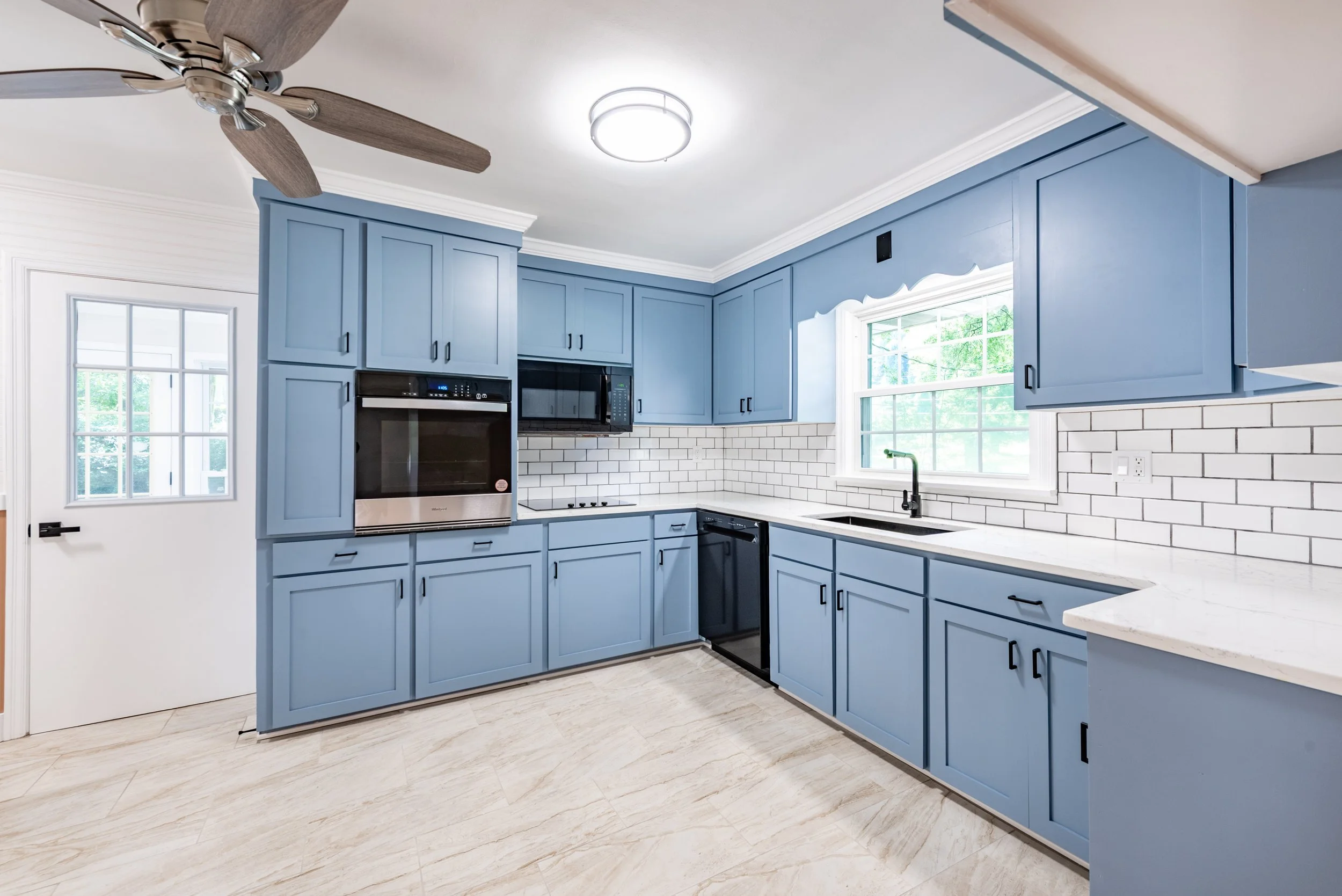 Modern kitchen with blue cabinets, white subway tile backsplash, black appliances, large windows, white ceiling with crown molding, and wooden flooring.