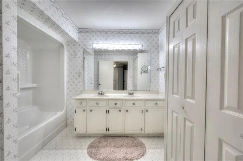 Bathroom with a large mirror, single sink vanity, and a bathtub with a shower. The walls have light-colored floral wallpaper, and there is a closet with double doors. A brown rug is on the white hexagonal tile floor.