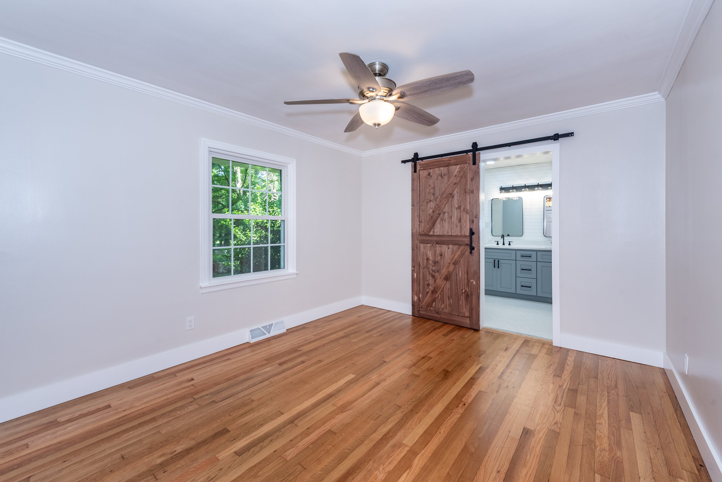 Empty room with hardwood floors, white walls, a window showing greenery outside, and a wooden sliding barn door leading to a bathroom with a vanity and mirror.