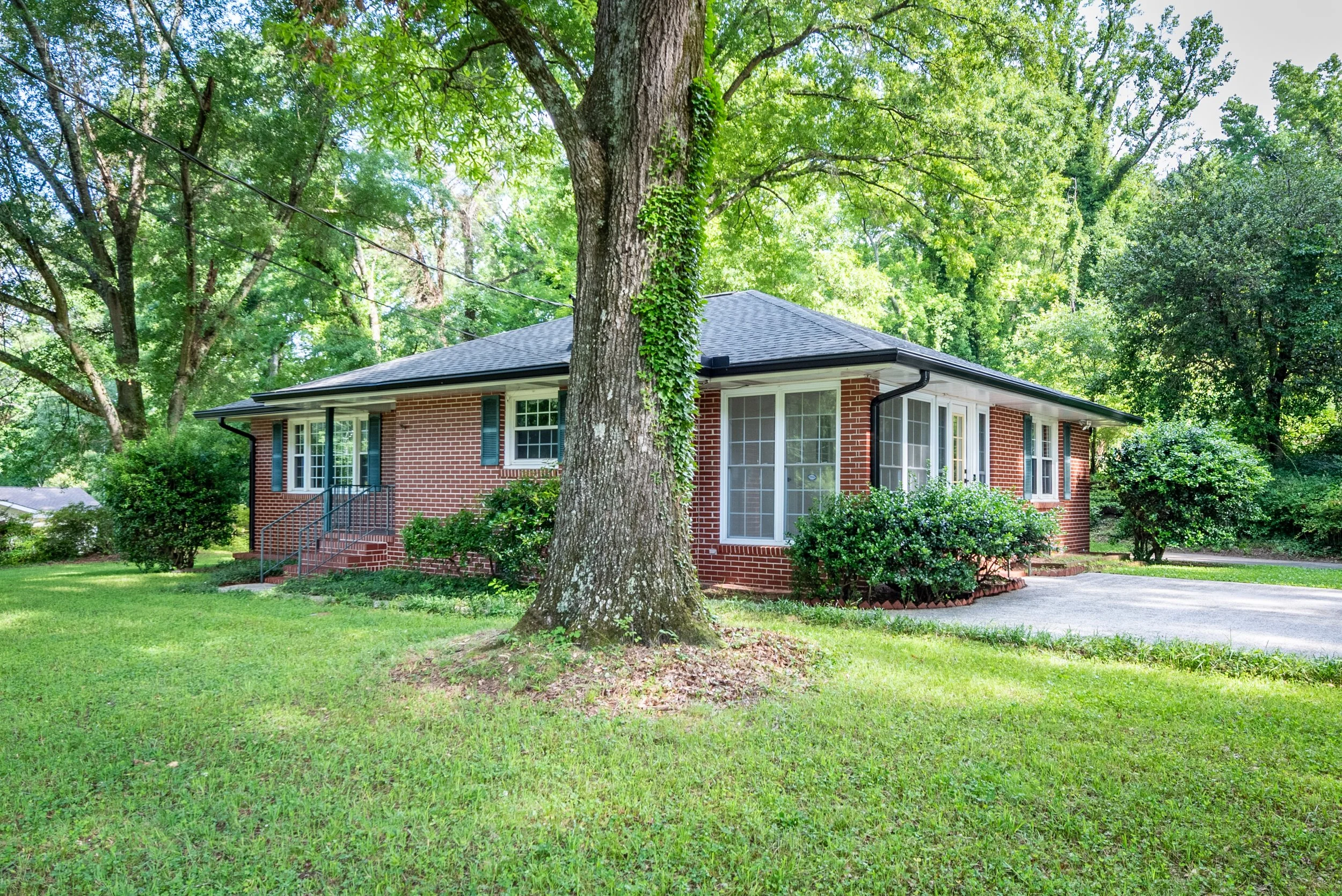 A brick house surrounded by green trees and a well-maintained lawn, with a large tree in front and a driveway on the right side.
