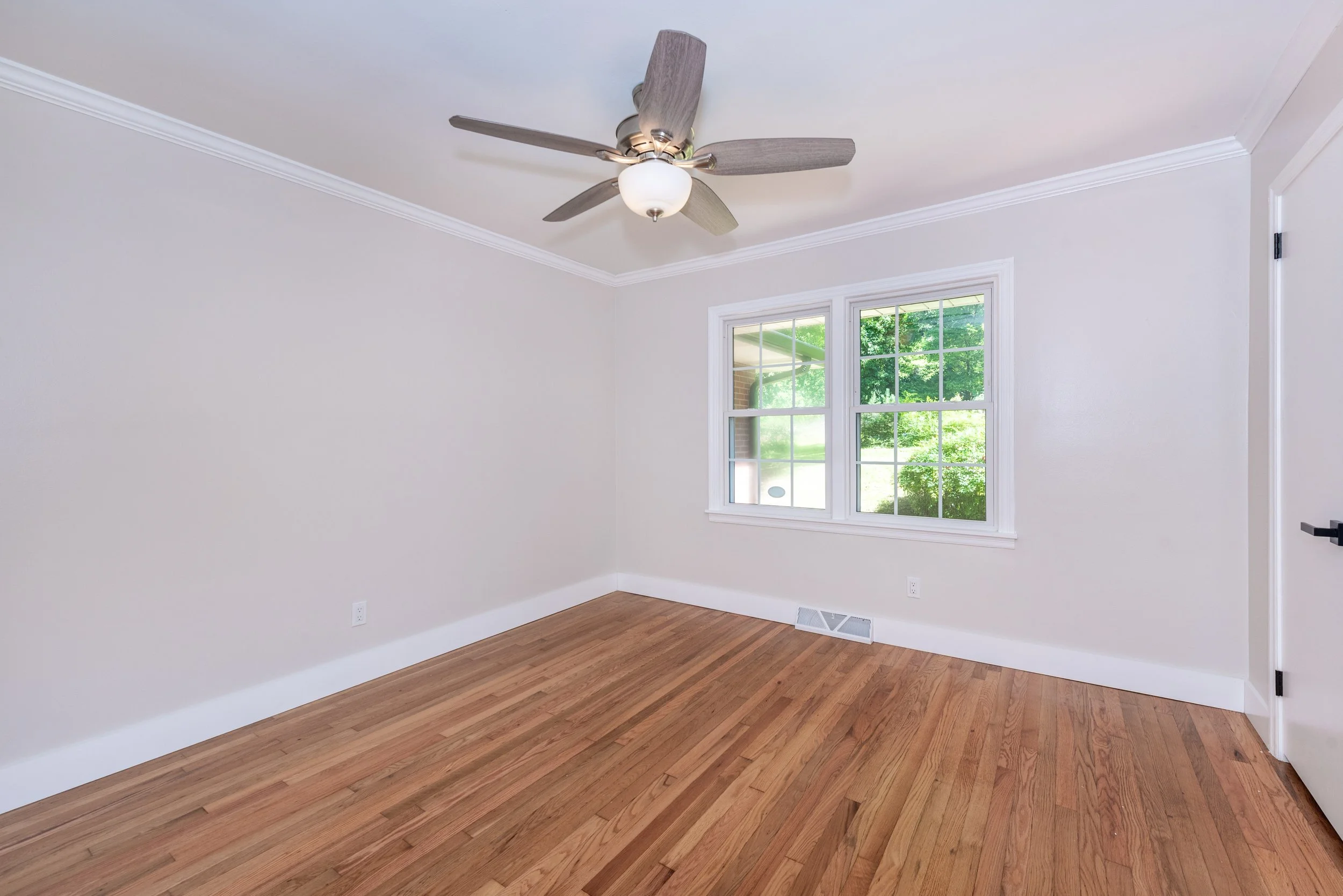 Empty room with hardwood floors, white walls, a ceiling fan with light, and a window showing green trees outside.