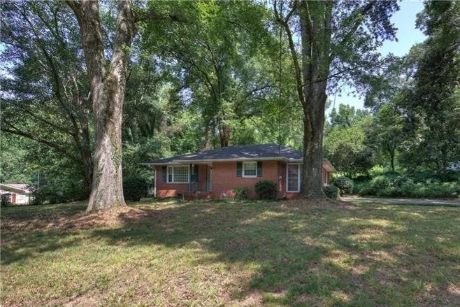 A single-story brick house surrounded by large trees in a grassy yard under a blue sky.
