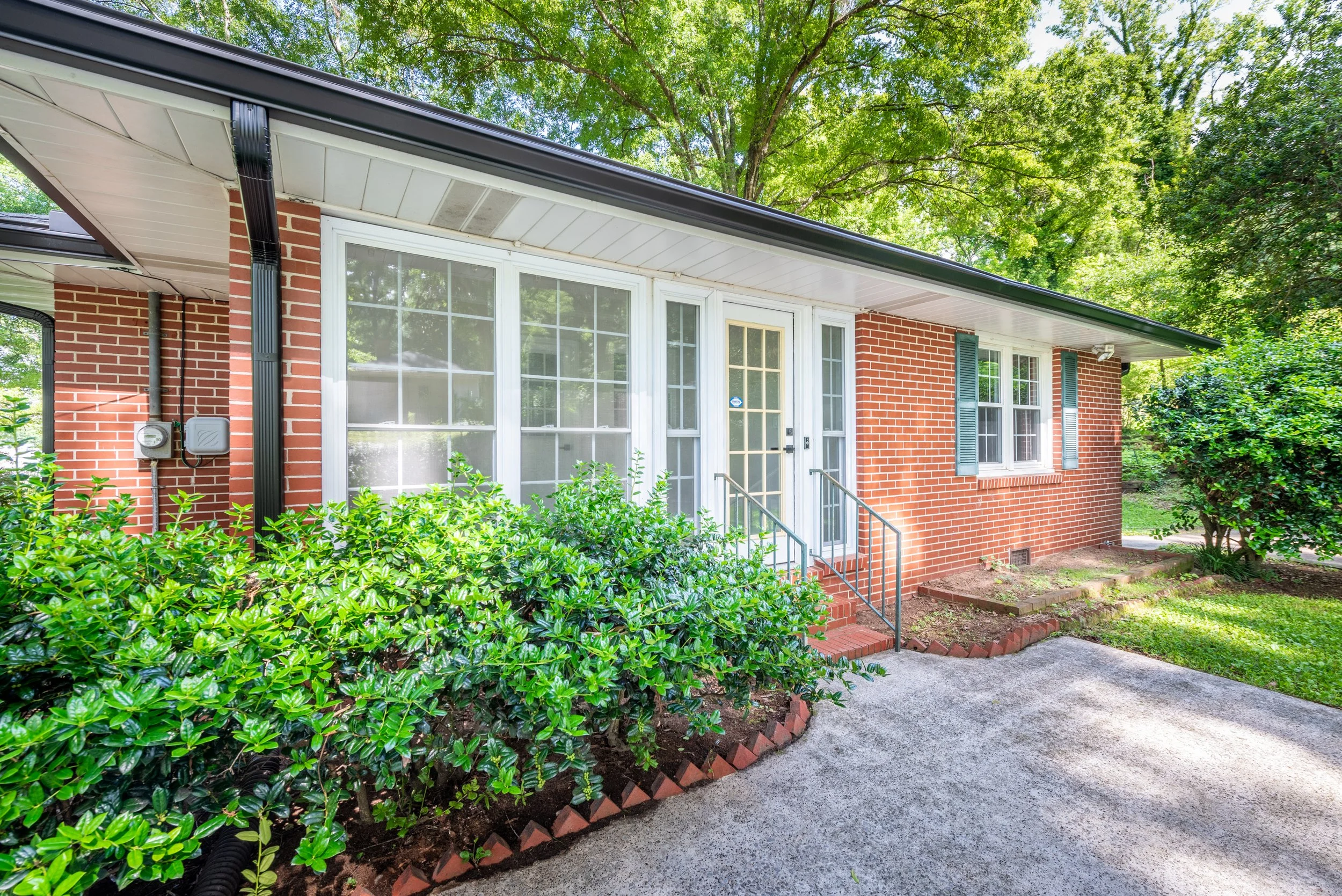 Red brick house with white trim, large windows, front porch, green bushes, and surrounded by tall green trees in a neighborhood.