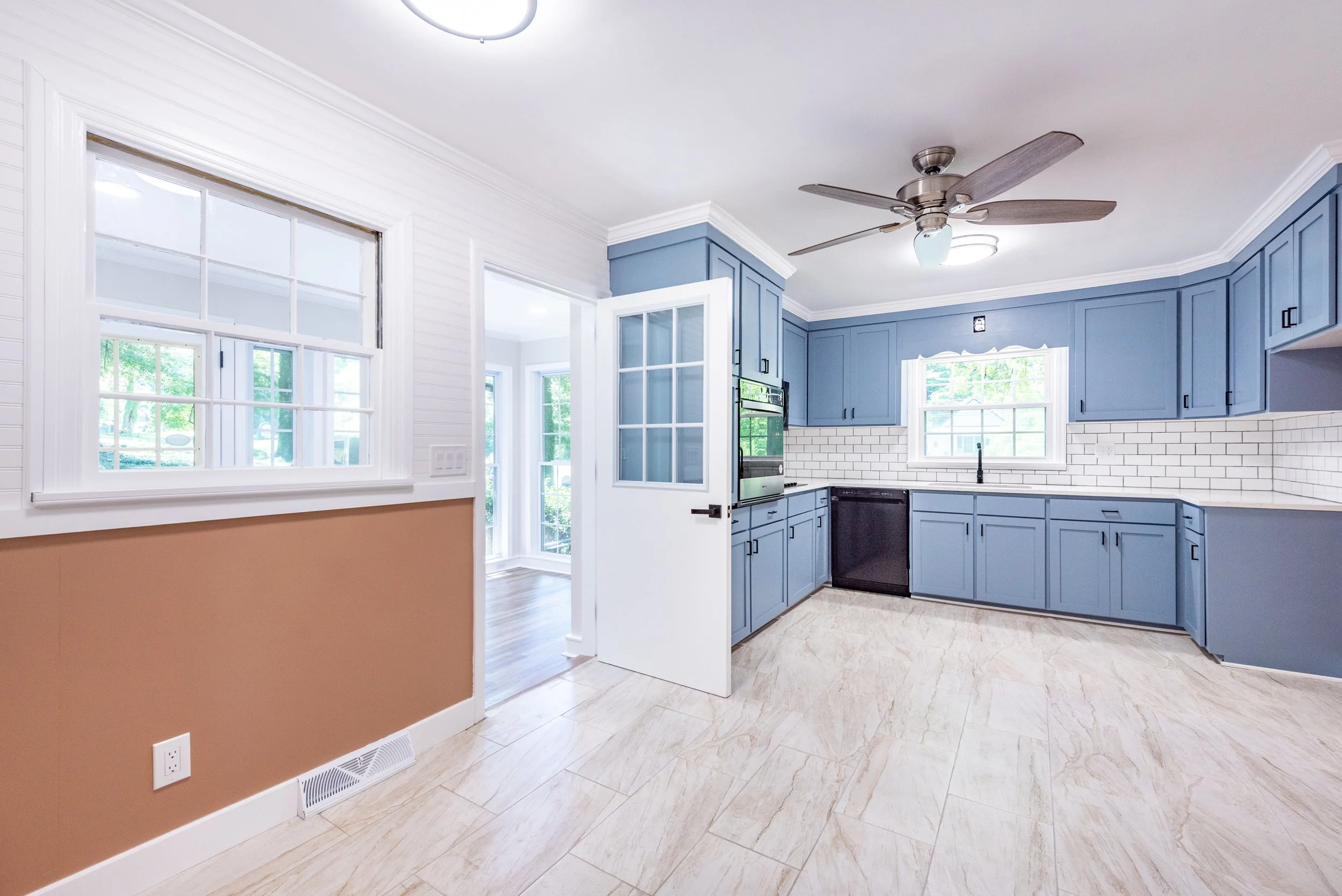 Kitchen with blue cabinets, white backsplash, large window, and a wooden floor in an open space.