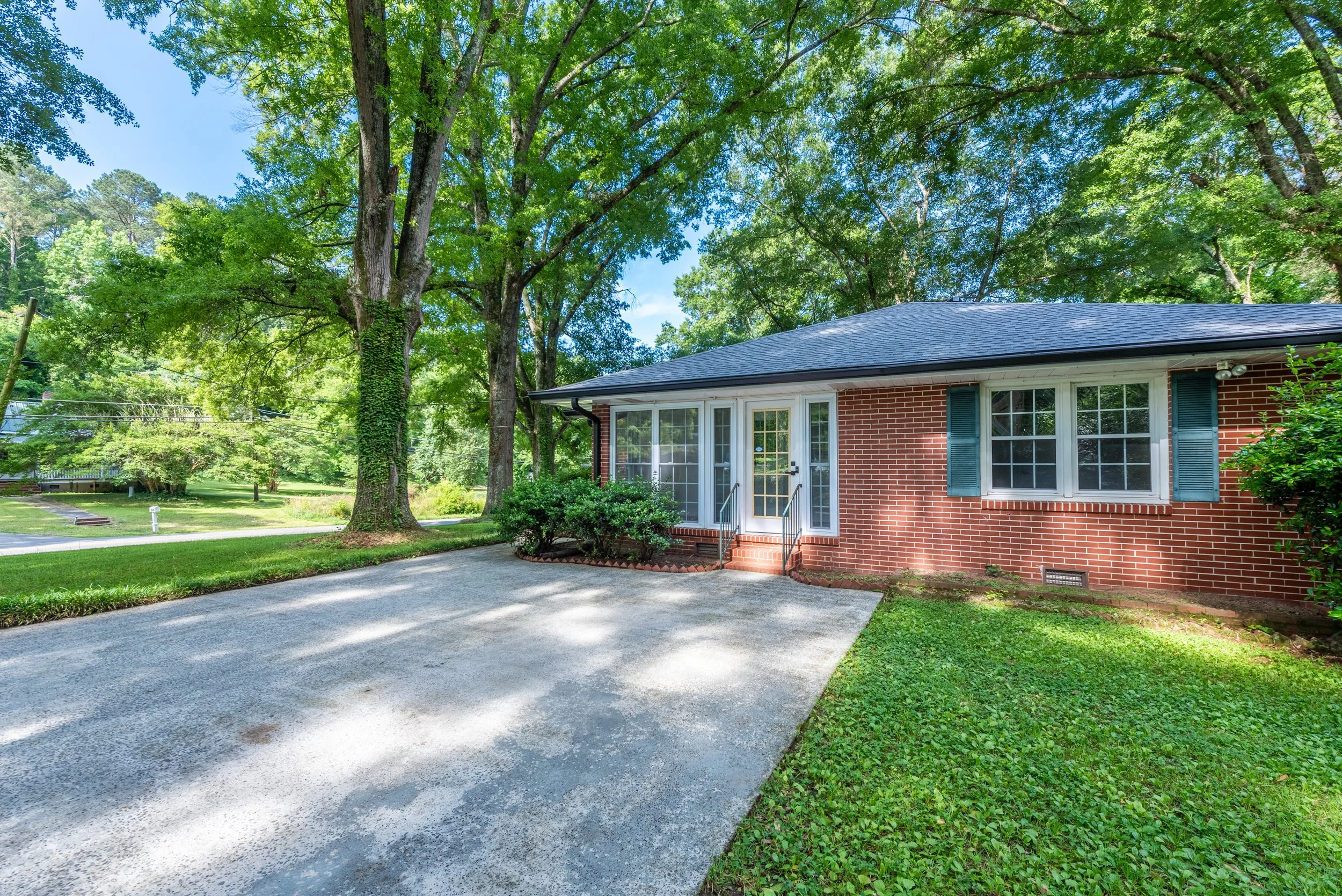 A brick house with a front porch, surrounded by green trees and grass, with a gravel driveway in front.