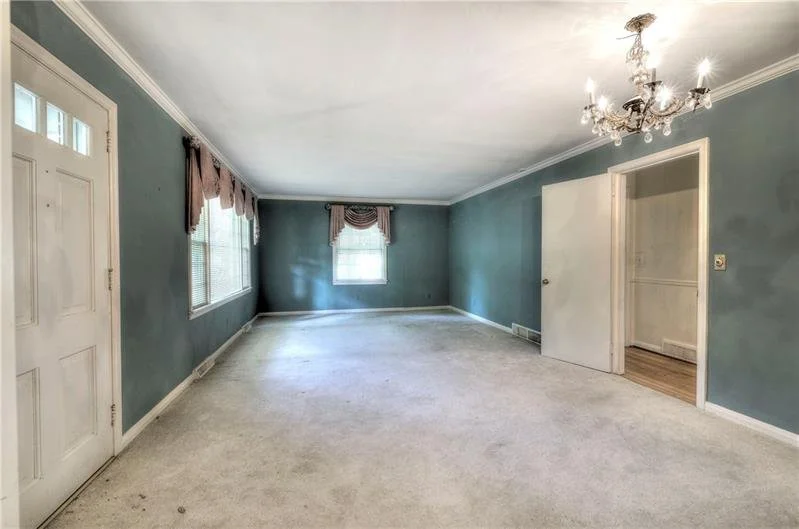 Empty living room with blue walls, white ceiling, chandelier, two windows with brown curtains, and a doorway leading to another room.