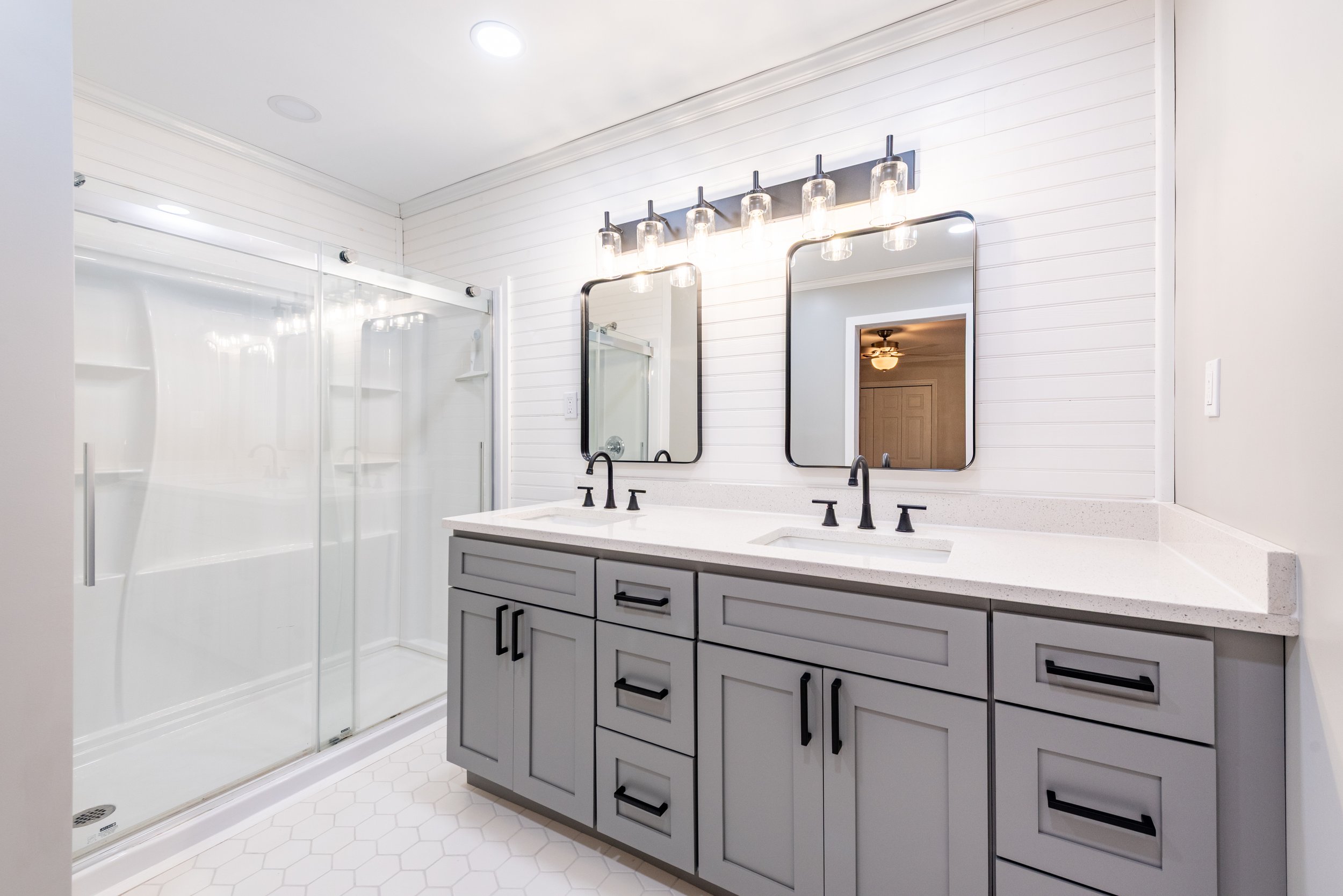 A modern bathroom with a grey vanity, black hardware, two mirrors, and a glass-enclosed shower with white walls and hexagonal tile flooring.