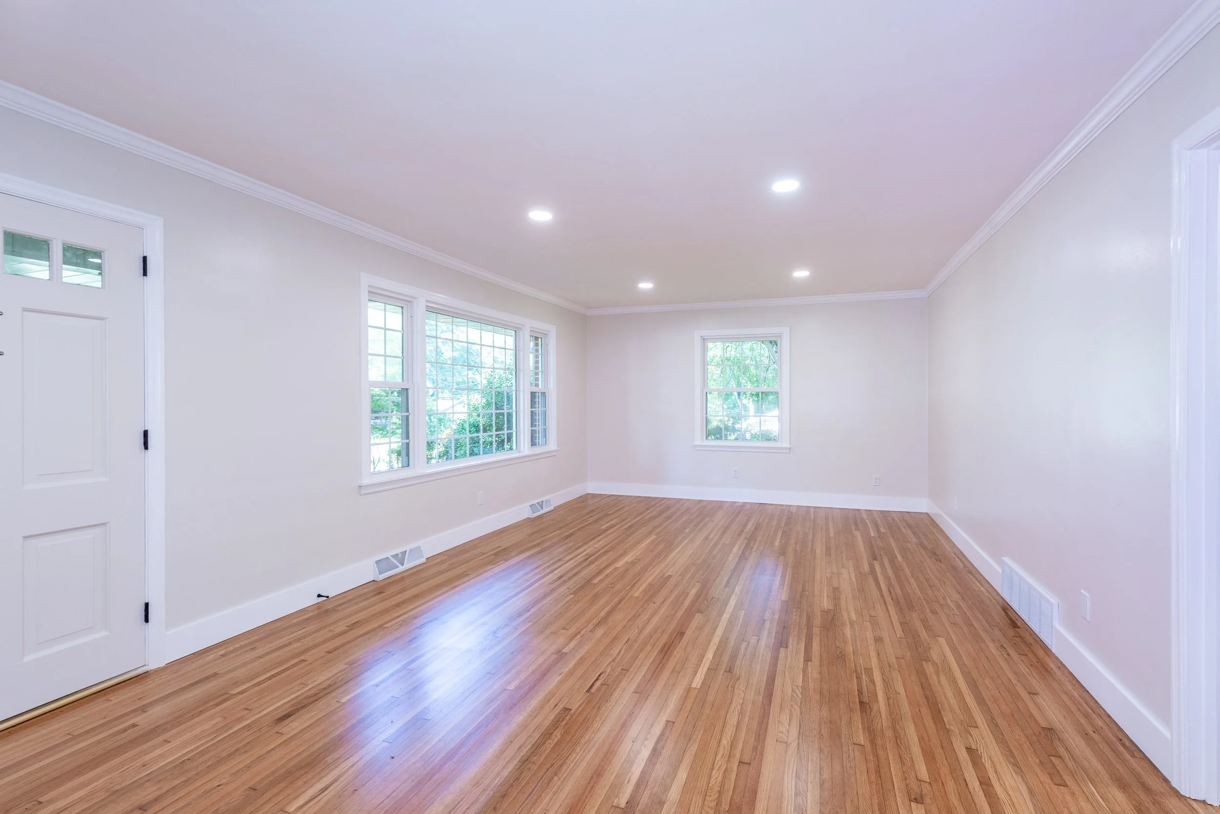 Empty living room with white walls, hardwood floors, and multiple windows letting in natural light.