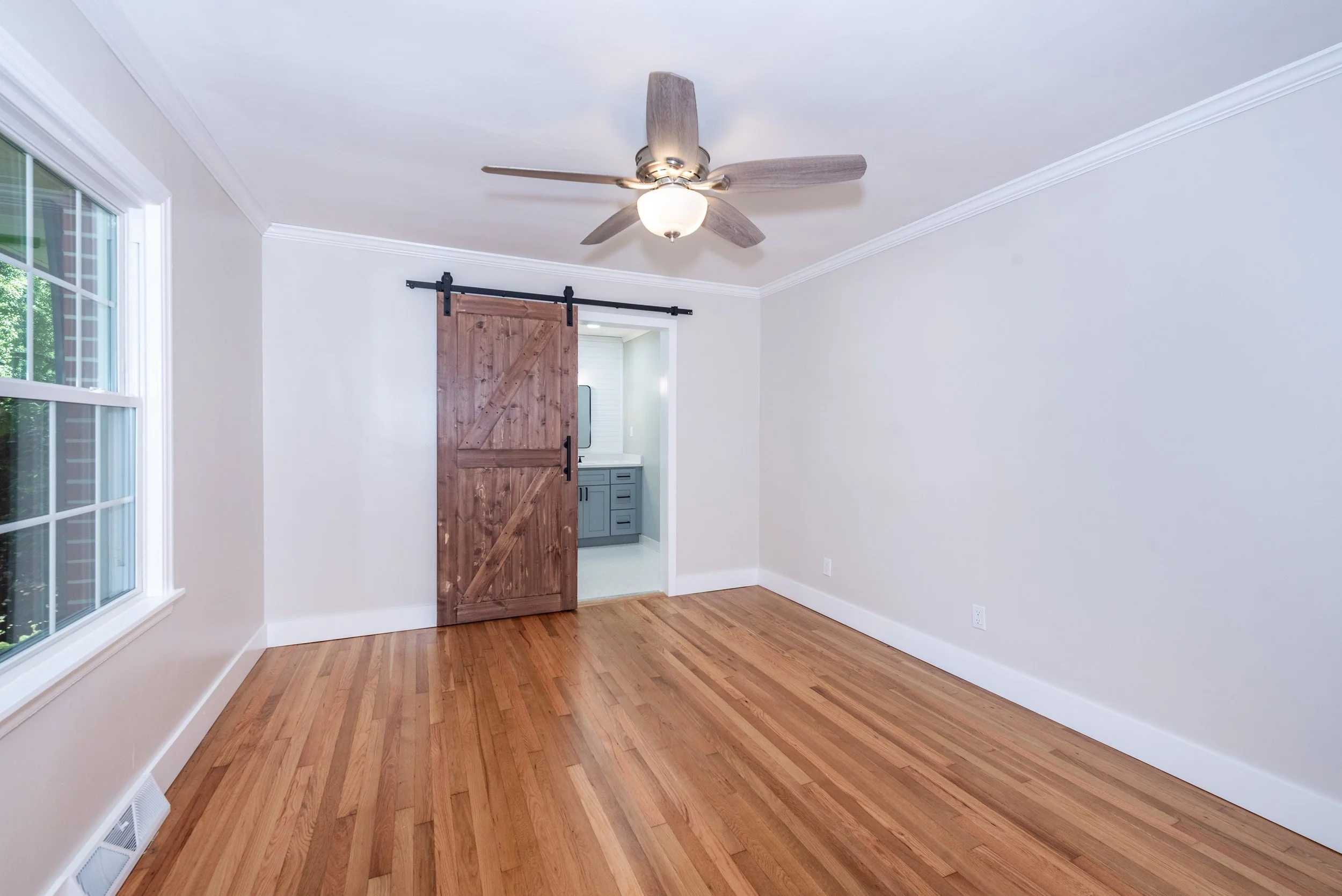 Empty room with hardwood flooring, a ceiling fan, a large window, and a wooden sliding door leading to another room.
