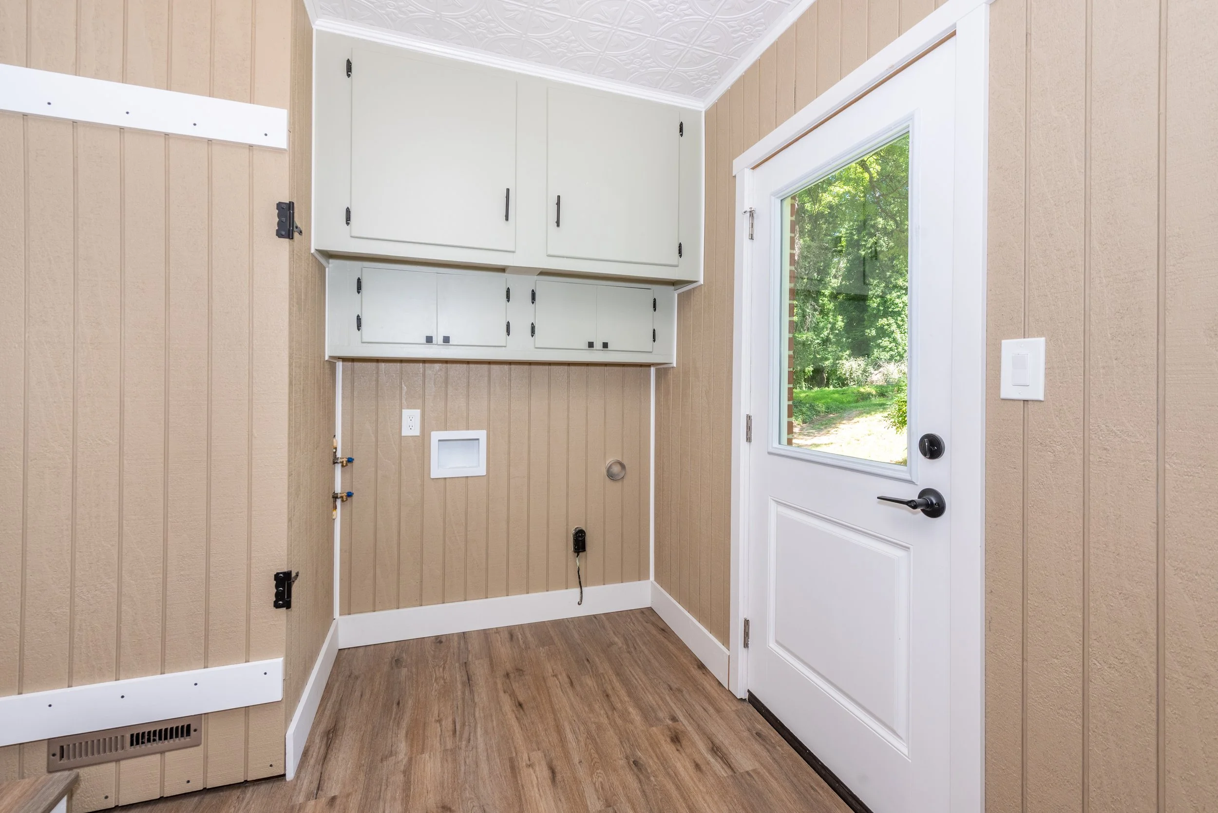 Interior view of a laundry area with beige wainscoted walls, white cabinets, a window showing green trees outside, and a door with a black handle.