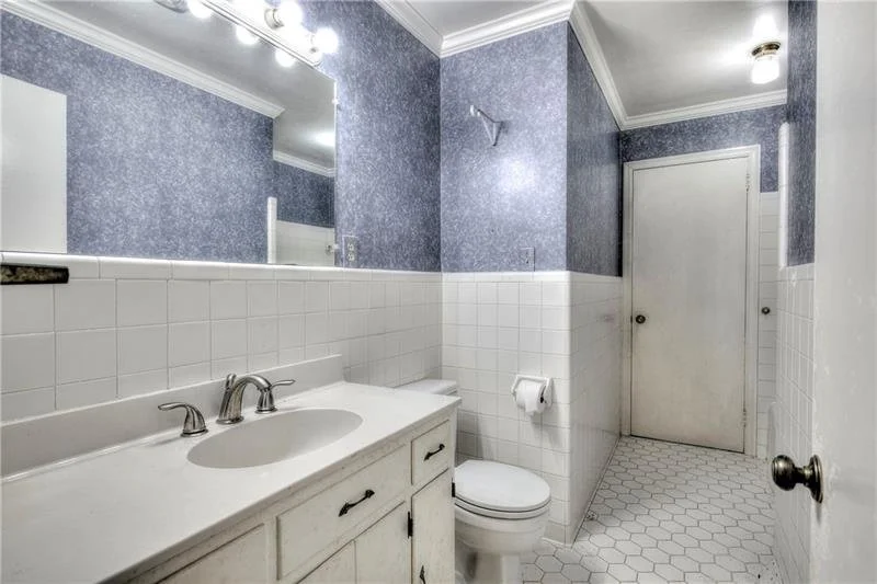 Small bathroom with blue textured walls, white tile halfway up, and hexagon floor tiles. Features a white vanity with sink, mirror, toilet, and door.