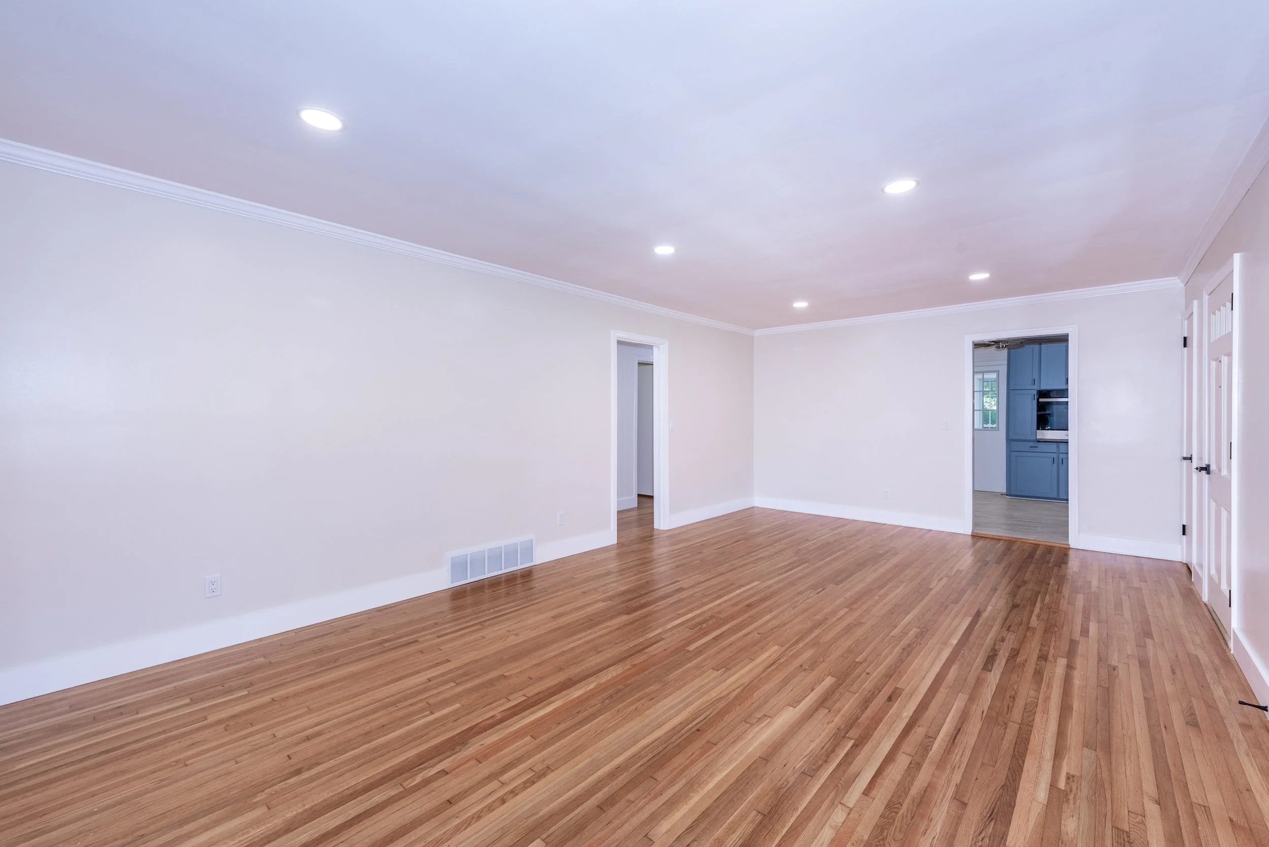 Empty living room with hardwood floors, white walls, and ceiling lights. Visible doorways leading to other rooms, including a kitchen with blue cabinets.
