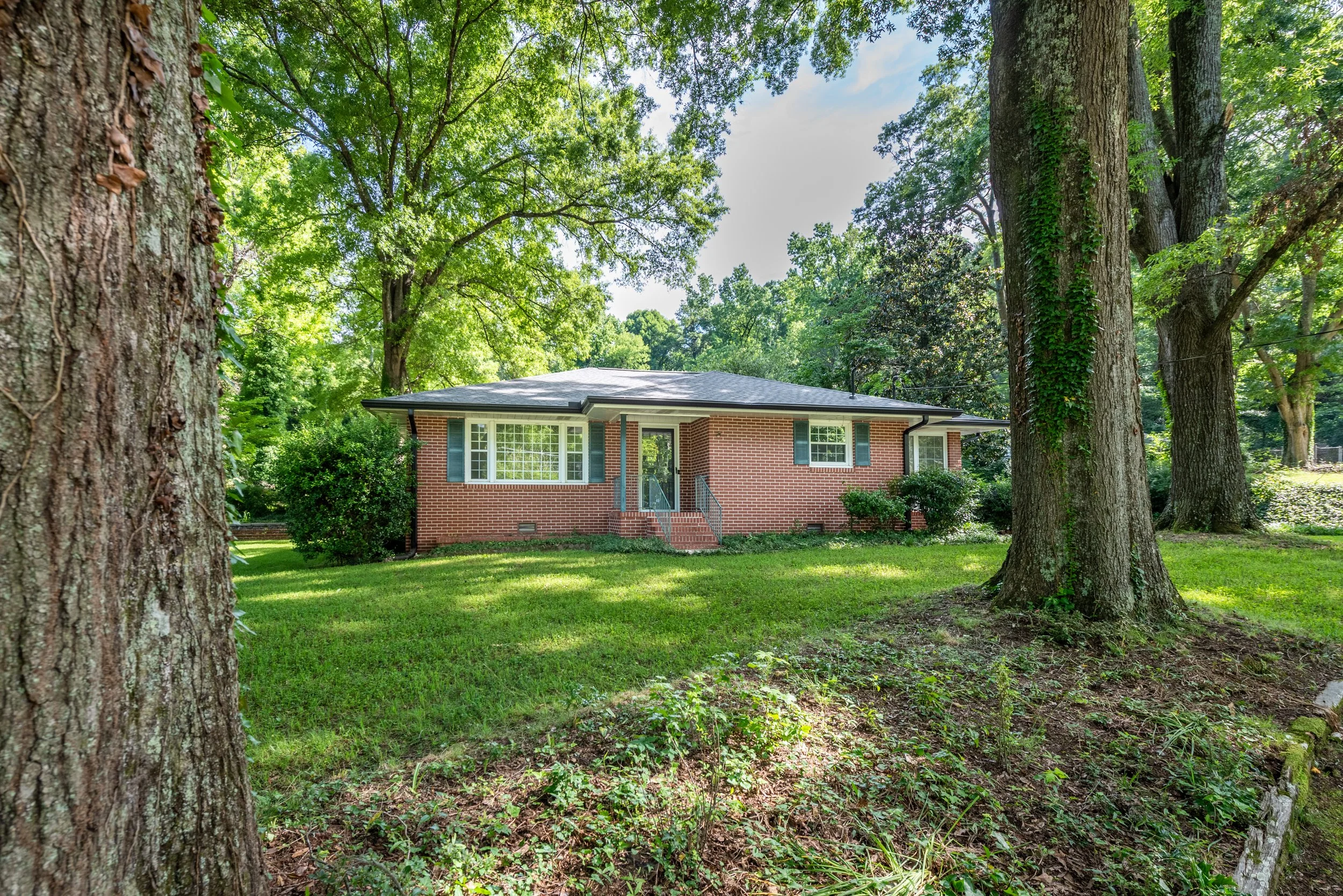 A brick house viewed through a wooded area with large trees and green foliage.