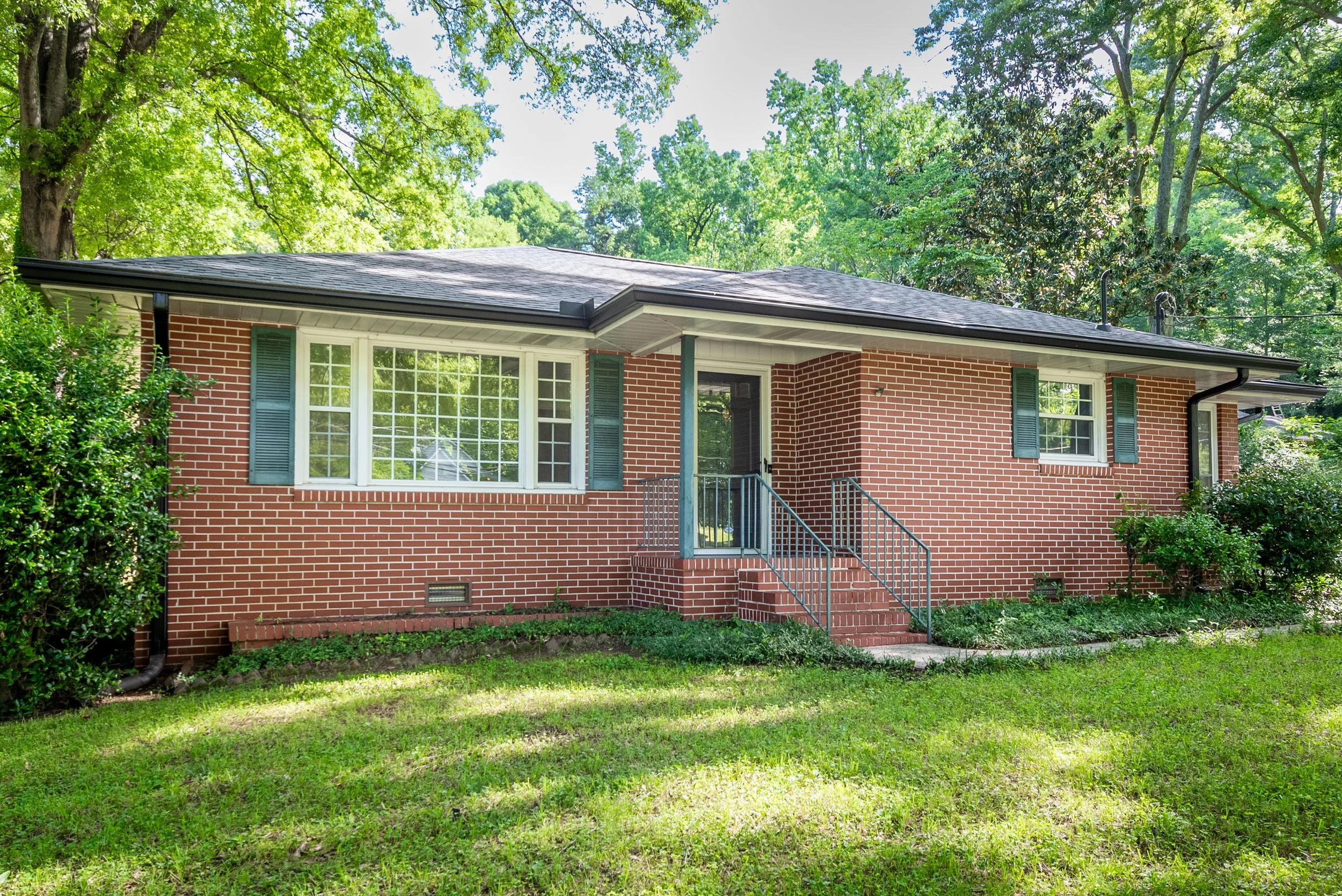 A red brick house with a gray shingle roof and white framed windows, surrounded by green trees and a well-maintained lawn.