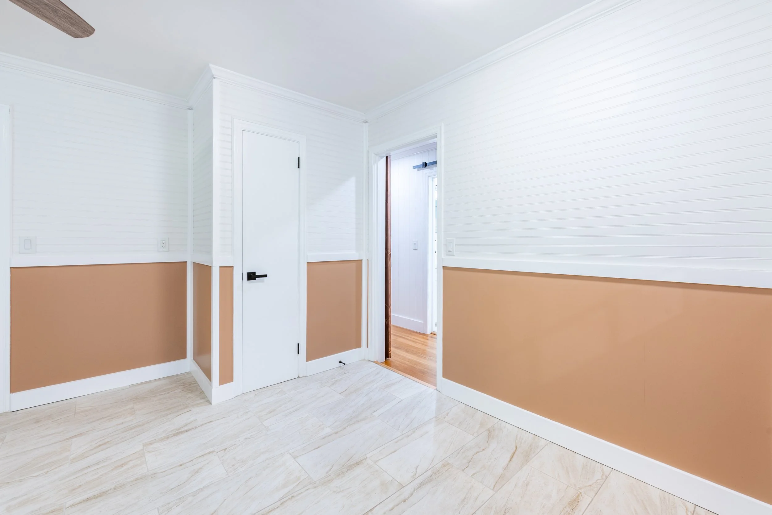 Interior view of a room with white walls featuring horizontal paneling, a two-tone color scheme with beige on the lower half, white trim, and a hardwood floor leading to another room.
