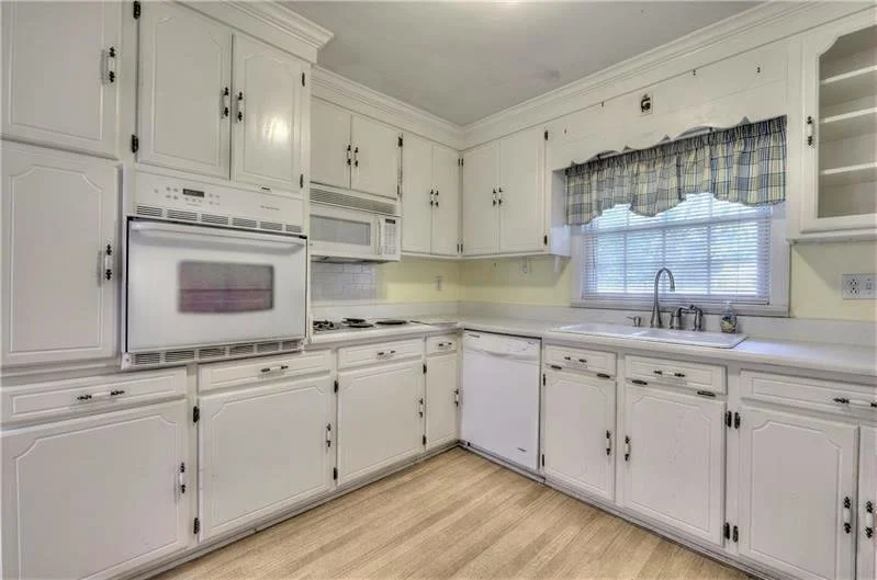 White kitchen with cabinets, a window with a blue and yellow checkered curtain, a double sink, and built-in appliances including an oven, microwave, and dishwasher, with light wood flooring.