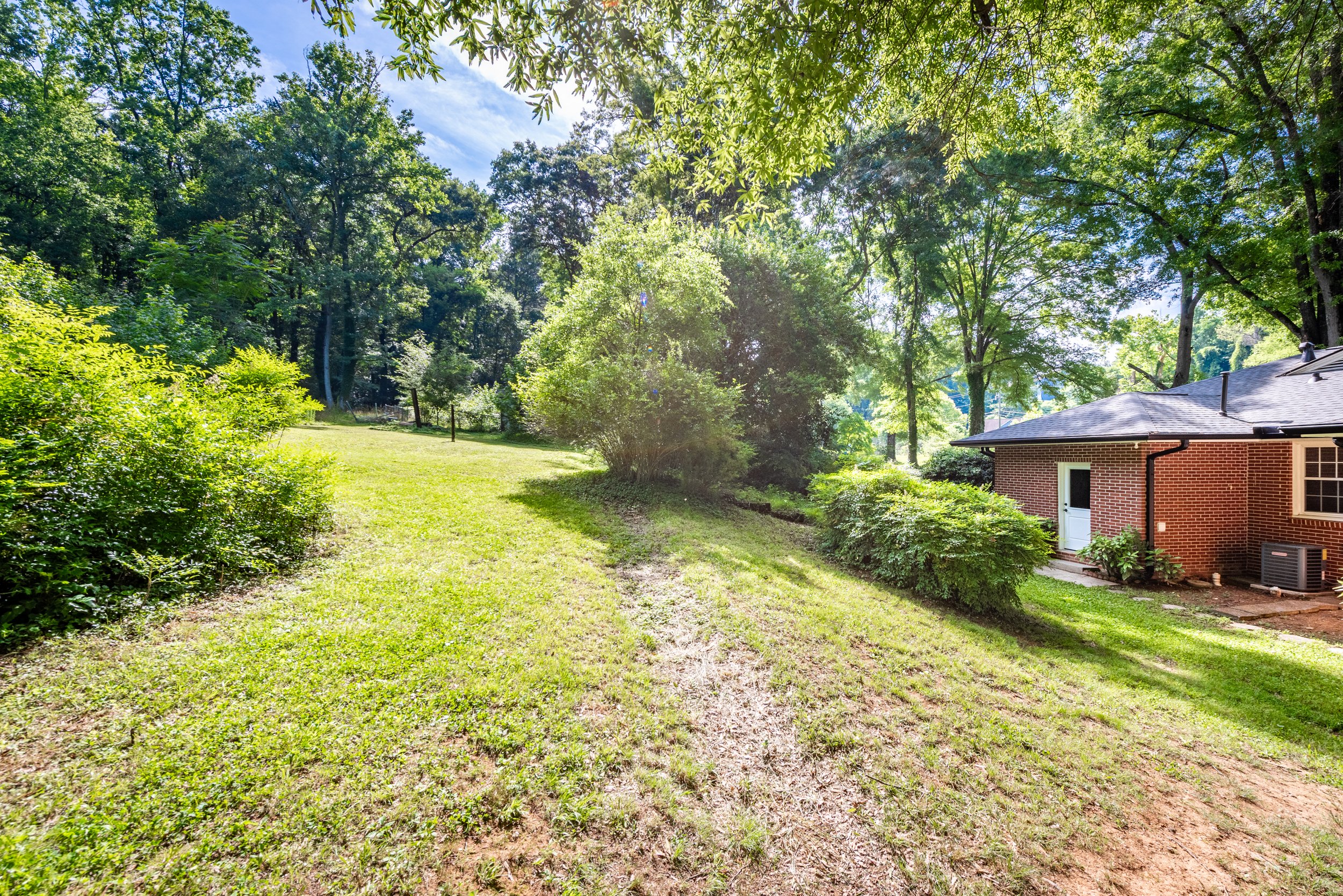 A backyard with green grass, bushes, and tall trees under a blue sky with some clouds. Part of a brick house with a white door and a gray roof is visible on the right side.