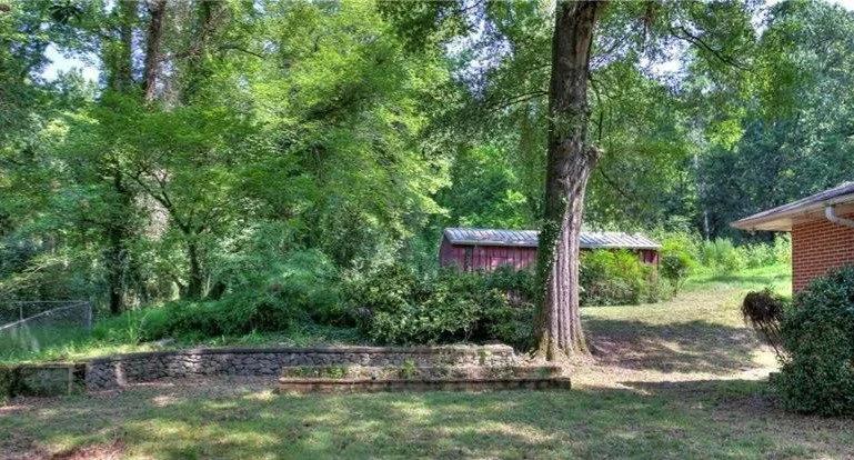 A backyard scene with green trees, a red shed, a brick house on the right, and a stone retaining wall.