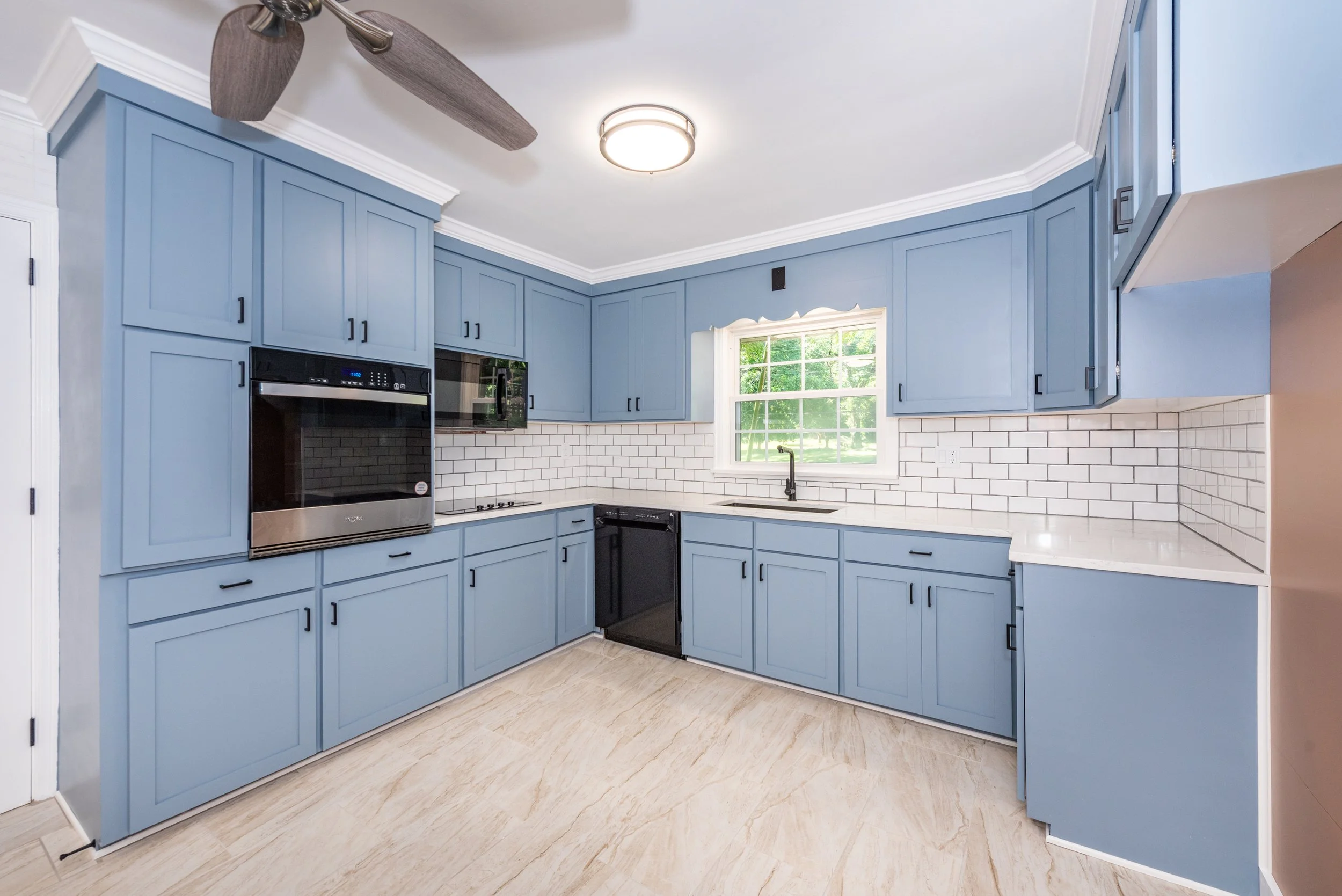 Bright blue kitchen cabinets with black hardware, white subway tile backsplash, a window with a view of trees, and a ceiling fan.
