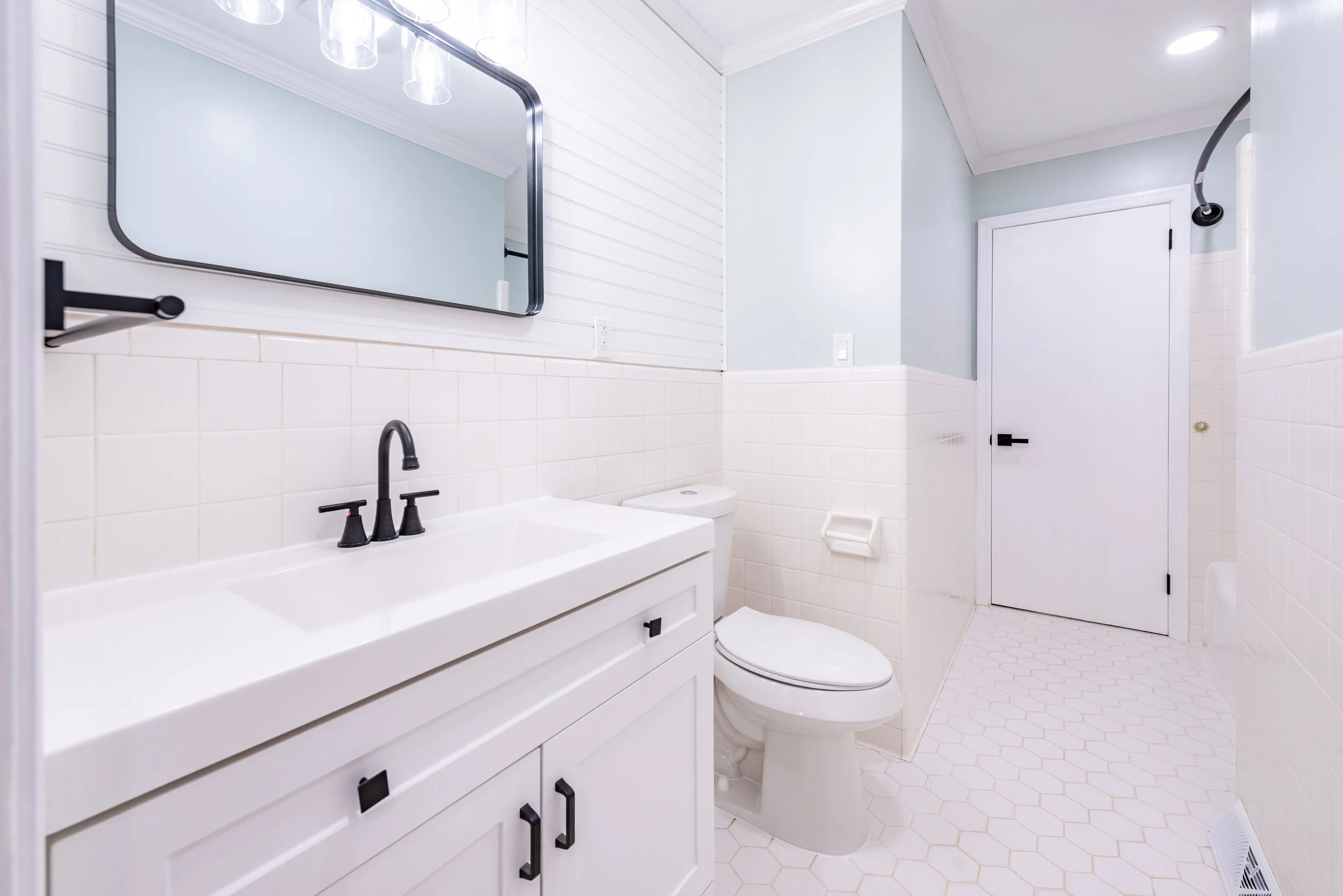 A bright modern bathroom with a white vanity, black cabinet handles, a large mirror, a black faucet, a toilet, and a partially visible shower with a glass door.