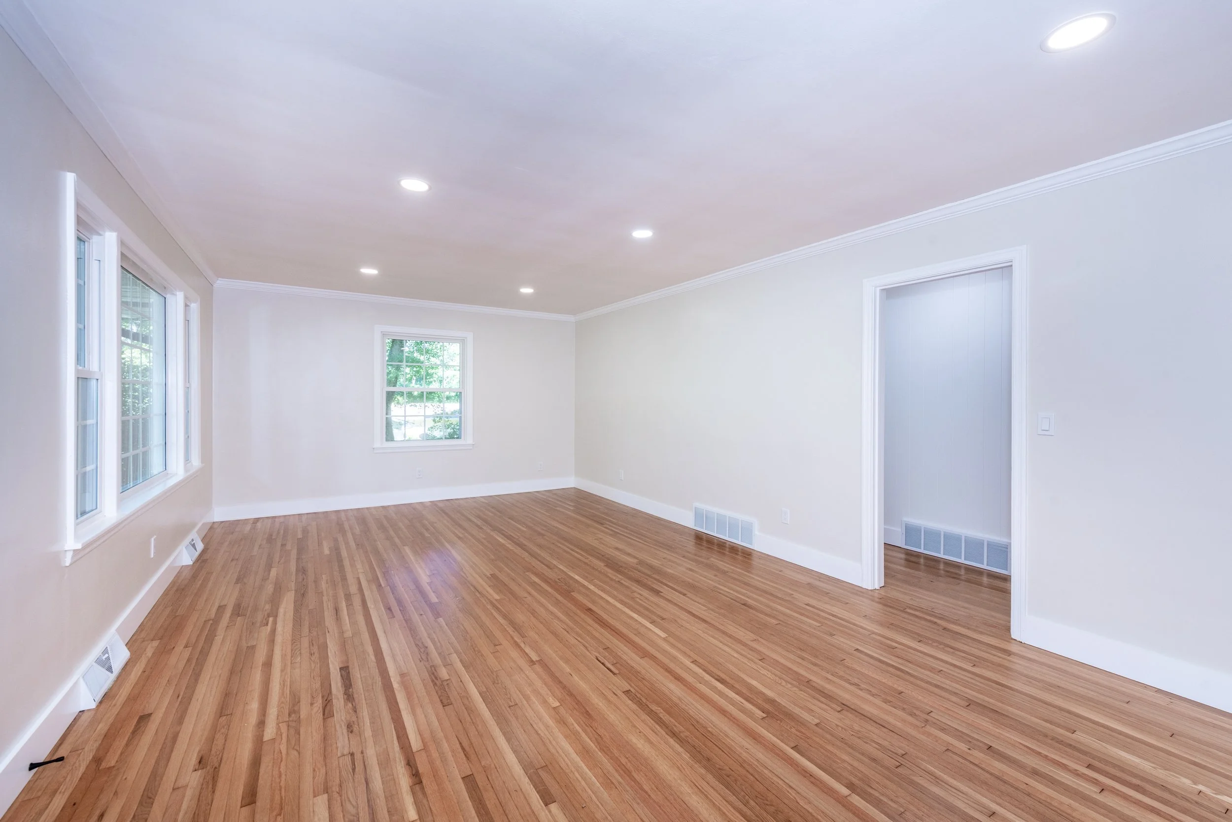 Empty living room with hardwood floors, white walls, multiple windows, and recessed ceiling lights.