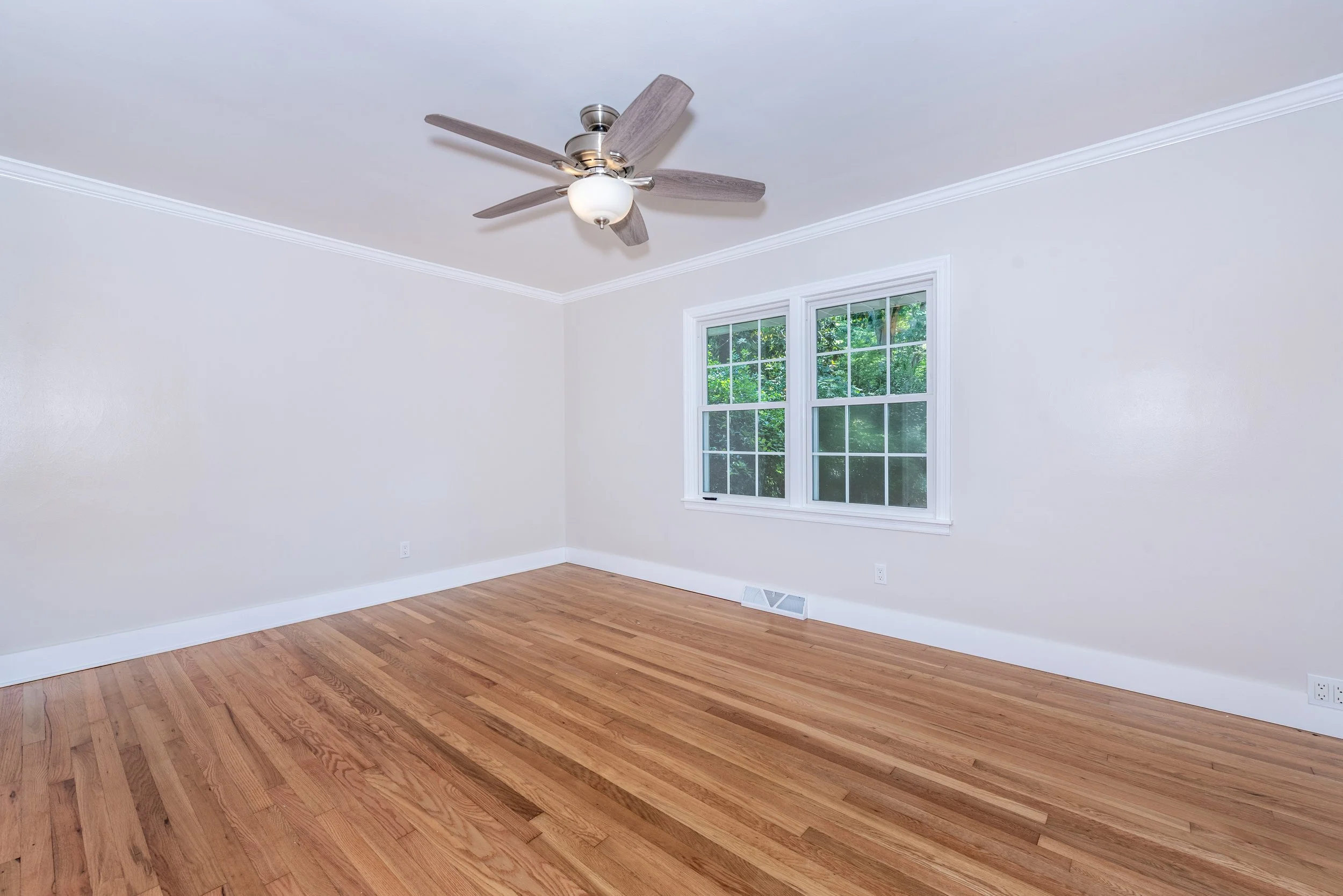 Empty room with hardwood floors, white walls, a ceiling fan with a light fixture, two windows with greenery outside, and baseboard vents.