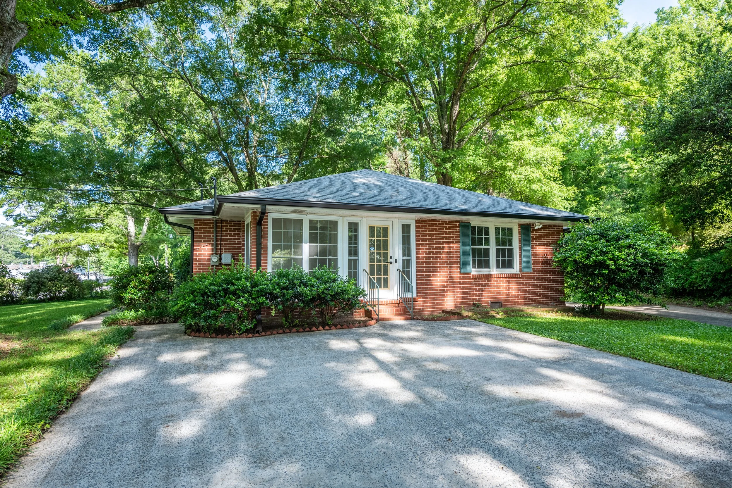 A brick house with white window frames, a yellow door, and green shutters, surrounded by lush trees and bushes, with a gravel driveway in front.