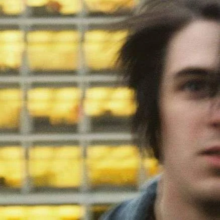 Close-up of a person with dark hair and pale skin, with a blurred background of yellow and gray objects.