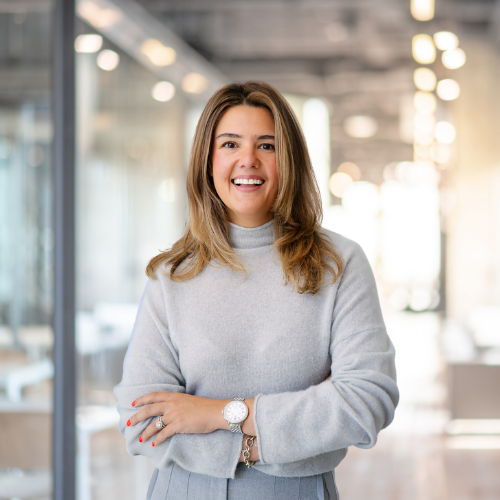 Woman with brown hair smiling, wearing a light gray sweater, in a modern office with glass walls and bright lighting.