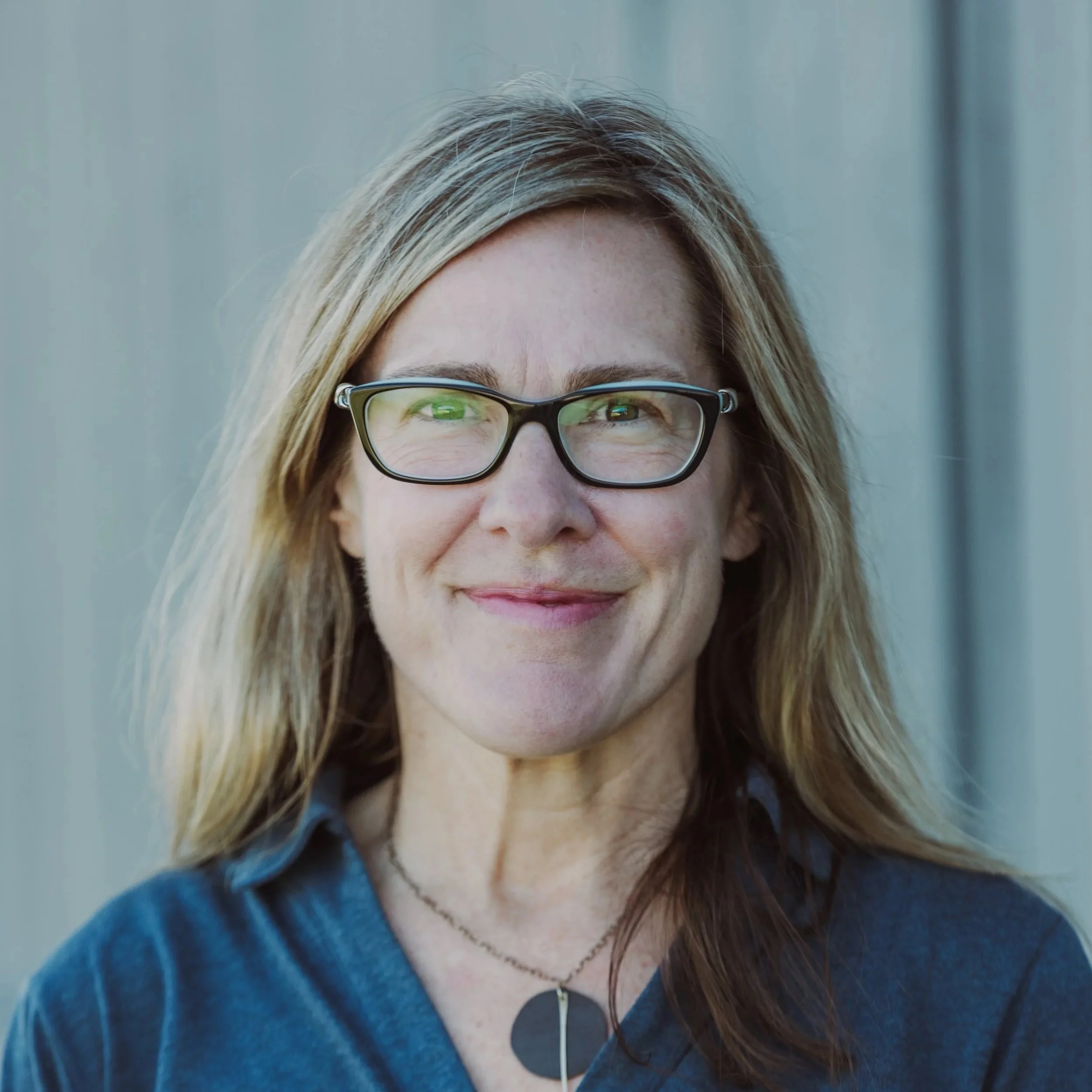 Close-up of a woman with long, wavy brunette hair wearing glasses, a blue top, and white earphones.