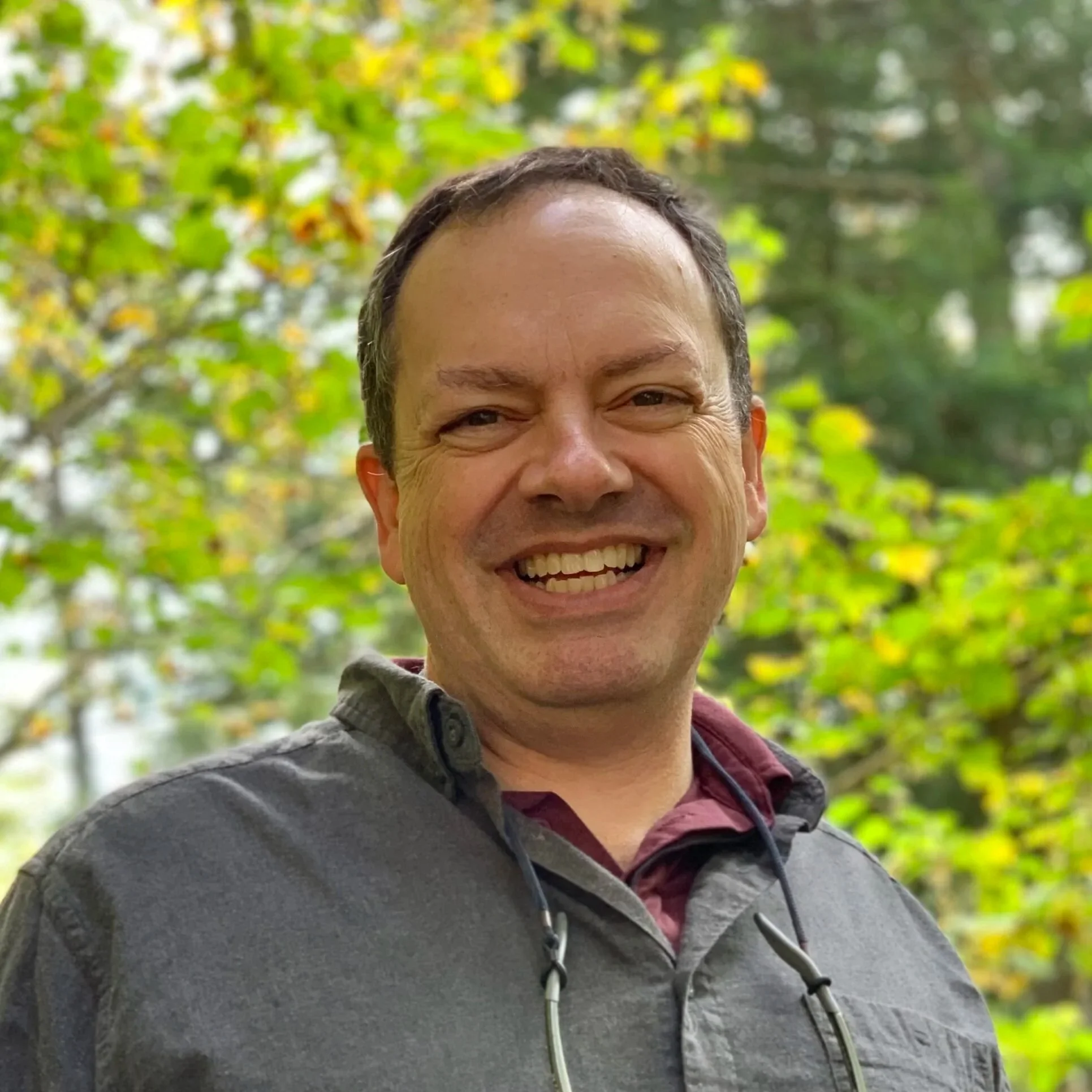Close-up of a smiling man with short brown hair, wearing a gray jacket and standing outdoors with green trees in the background.