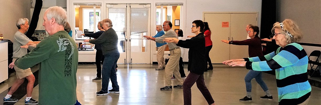 Group of elderly people participating in a tai chi class indoors, all with arms extended forward in a synchronized pose.