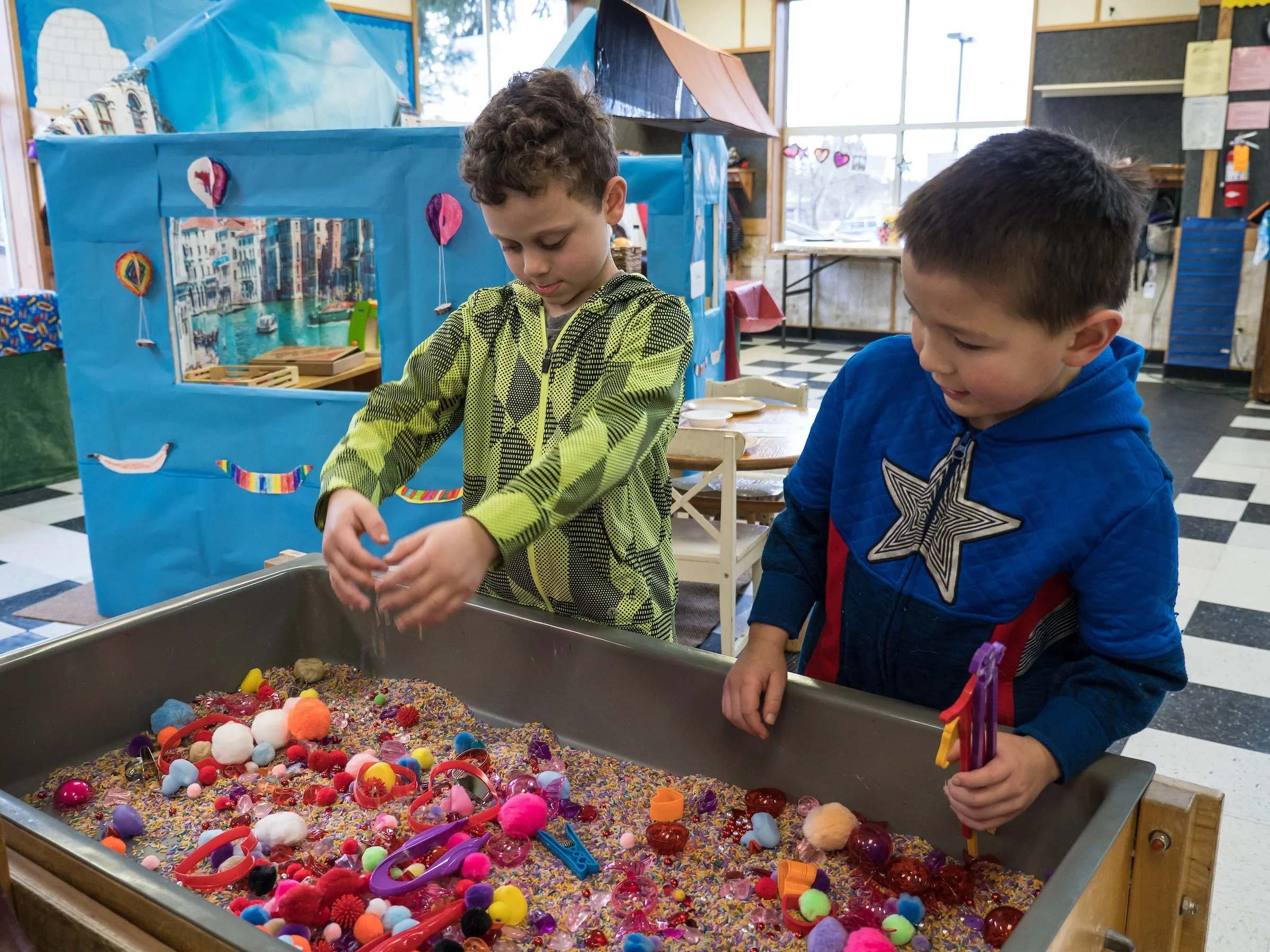 Two young boys are playing at a sensory bin filled with colorful small toys, pom-poms, and sprinkles in a classroom. One boy is wearing a yellow-green jacket, and the other is in a blue jacket with a star design. There is a blue show-and-tell display and large windows in the background.