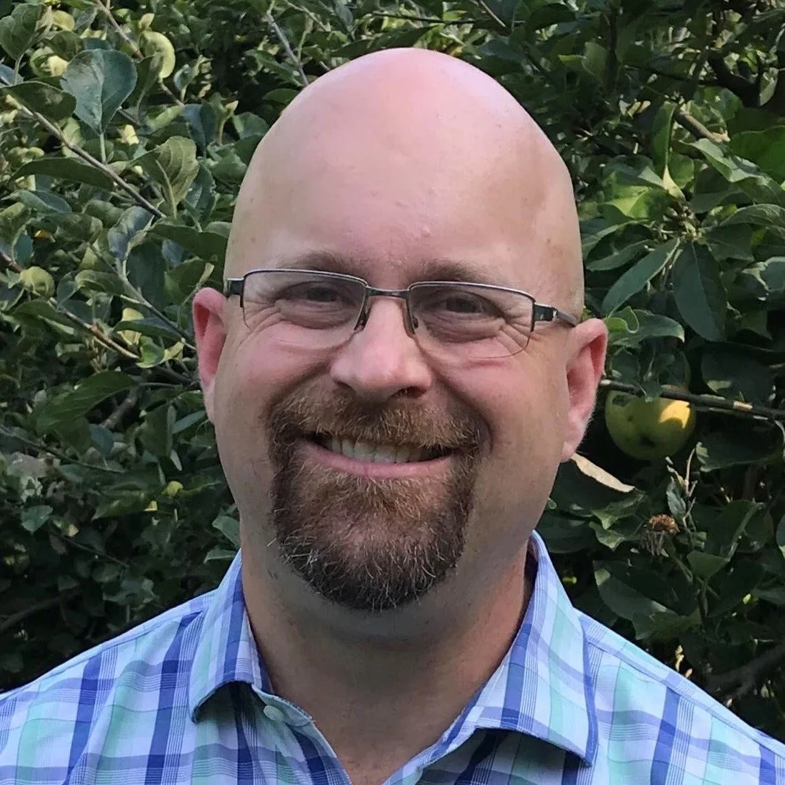 A bald man with glasses and a goatee, smiling, standing in front of green foliage and apples.