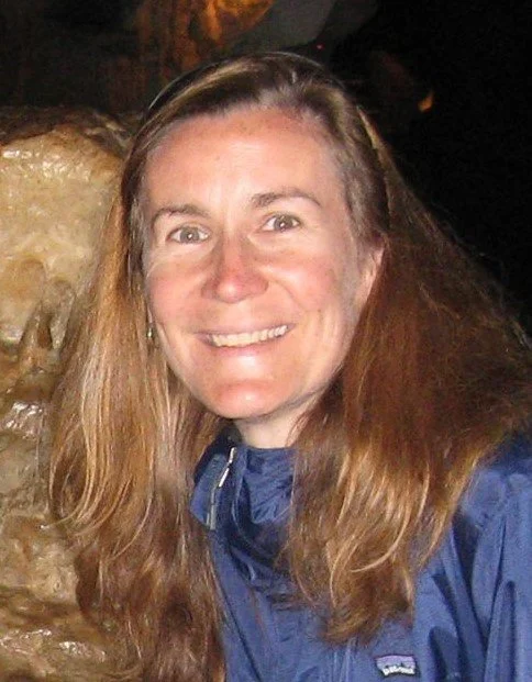 A woman with long brown hair smiling in front of a rocky background, wearing a blue jacket.