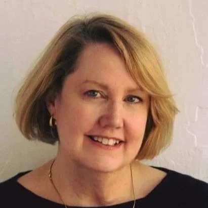 Portrait of a woman with shoulder-length blonde hair, wearing a black top, gold earrings, and a gold necklace, smiling at the camera against a white brick wall background.