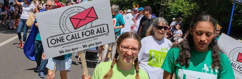 A group of people participating in a community event or walk, holding a sign that reads 'One Call for All' with a logo, on a sunny day.