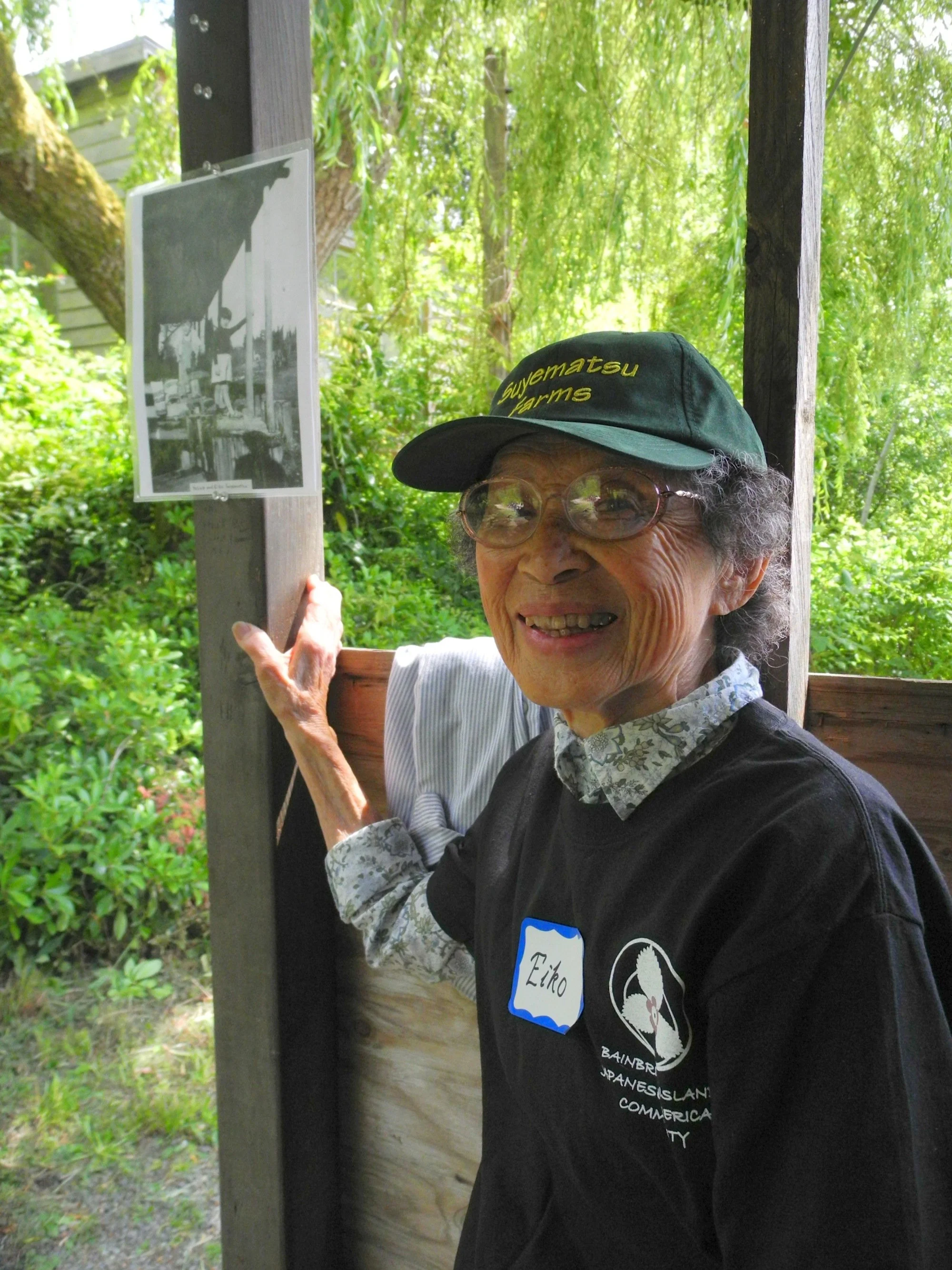 An elderly woman with glasses and a dark cap is smiling, holding a photo or sign on a wooden post in an outdoor green setting. She is wearing a black vest with a name tag that says 'Eiko' and an embroidered swan logo that reads 'Bainbridge Japanese I