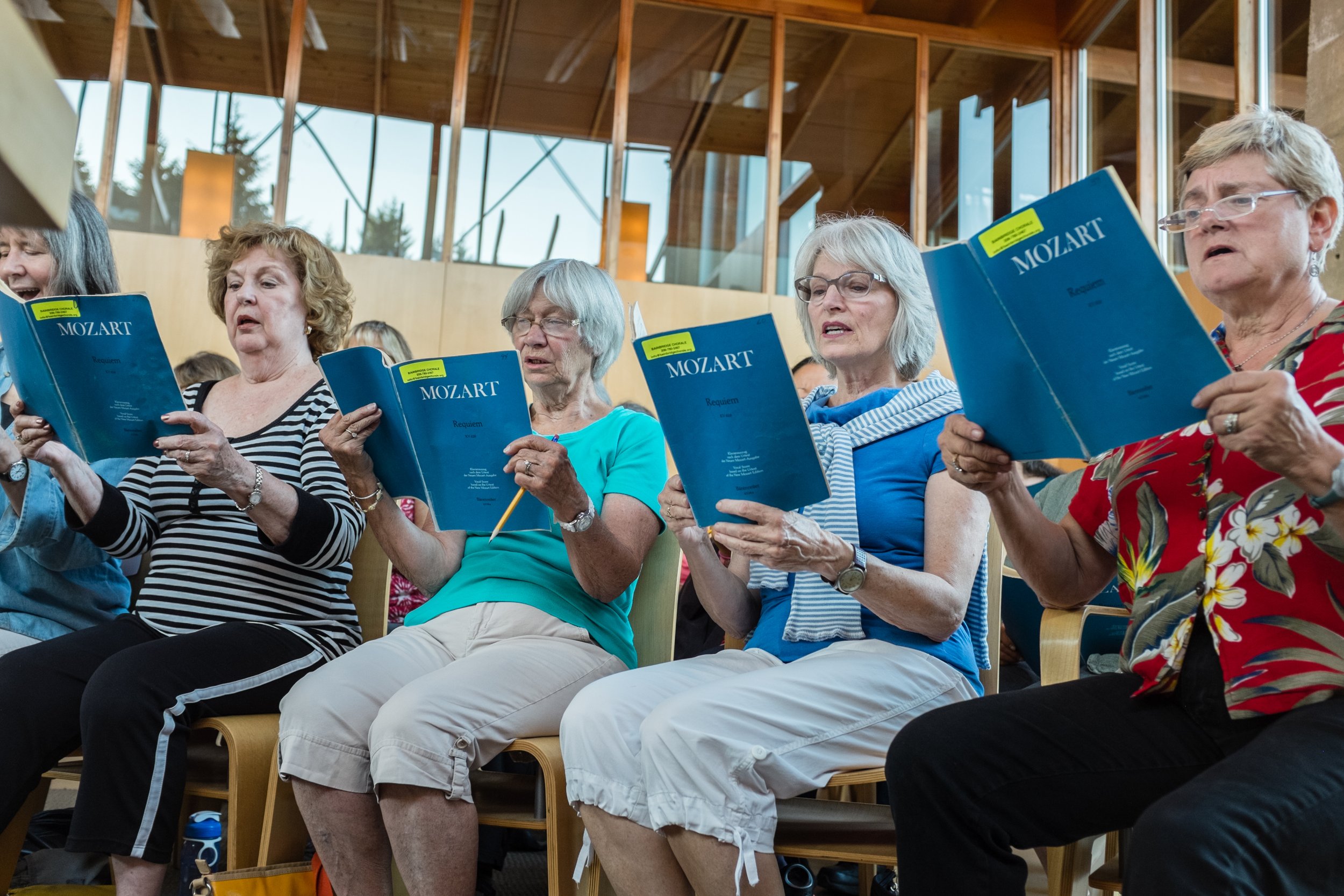Senior women singing from music books at a choir rehearsal indoors.